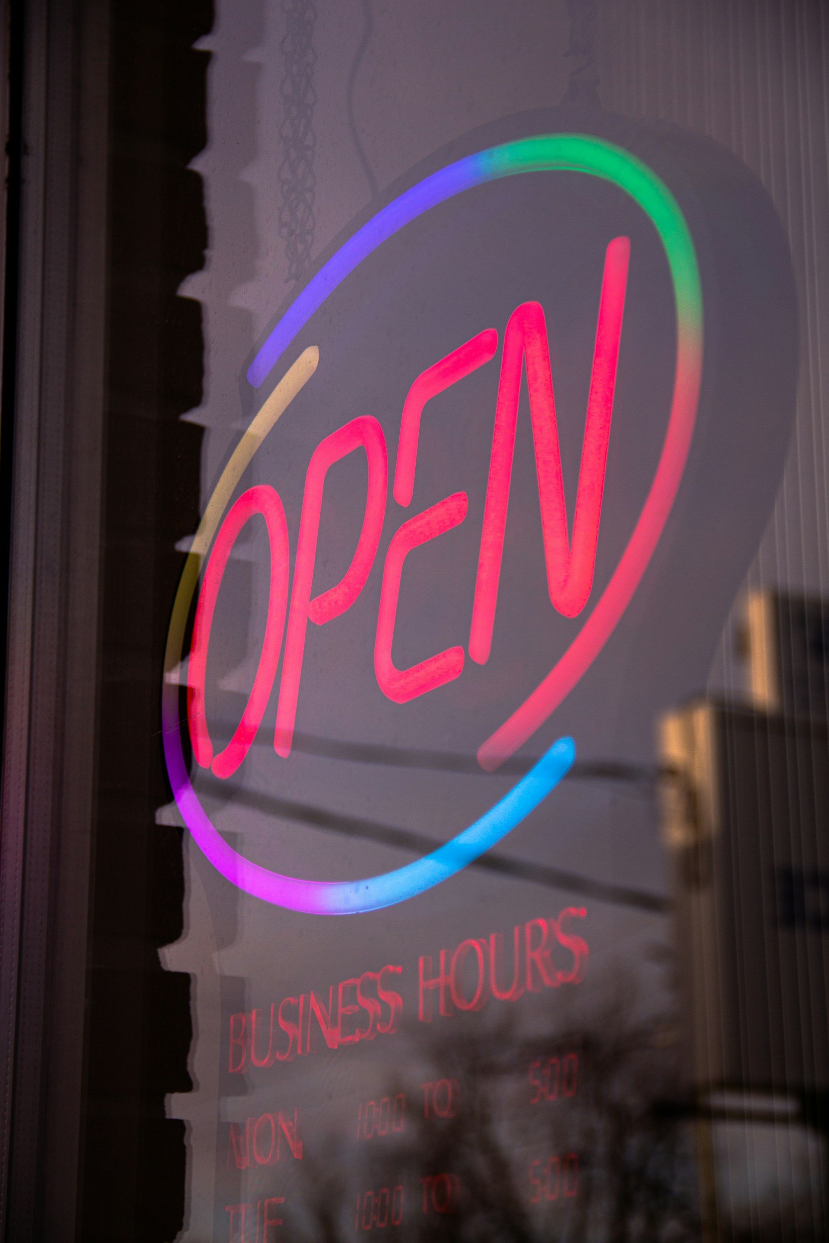 Neon sign that says "OPEN" in pink letters with colorful arcs around it, and below that, the business hours are listed in red on a dark background.