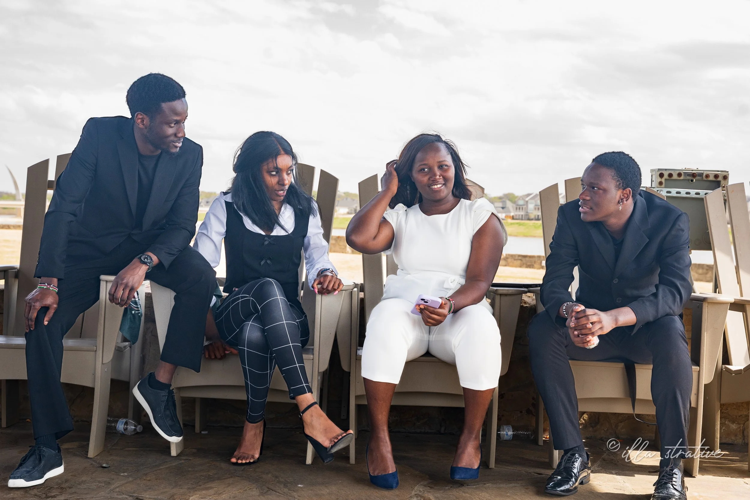 Four young adults sitting and talking outdoors on a cloudy day, with one woman dressed in white and a man in black suits, engaging in conversation.
