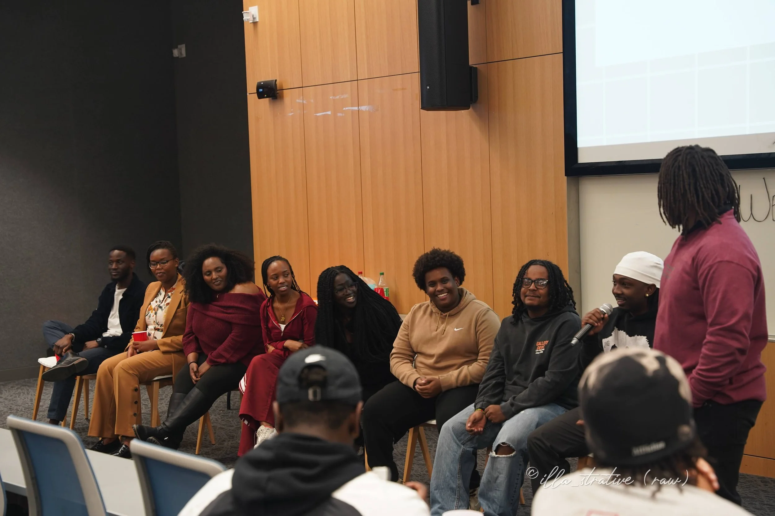 A panel of nine diverse people seated on chairs in a room, participating in a discussion or presentation, with one person standing and speaking into a microphone.