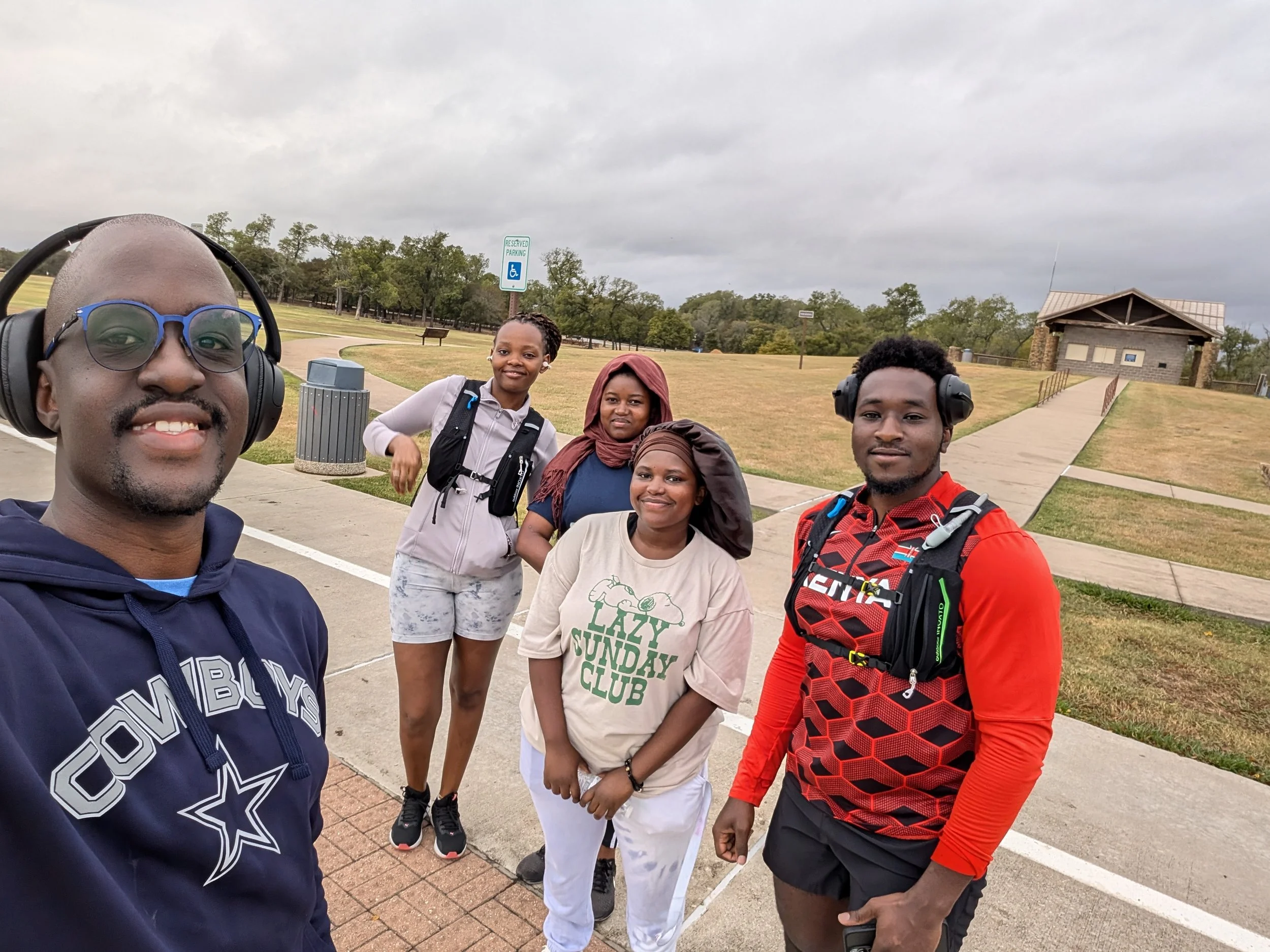 Group of five people with backpacks and headphones taking a selfie outdoors on a cloudy day, walking trail and a small building in the background.