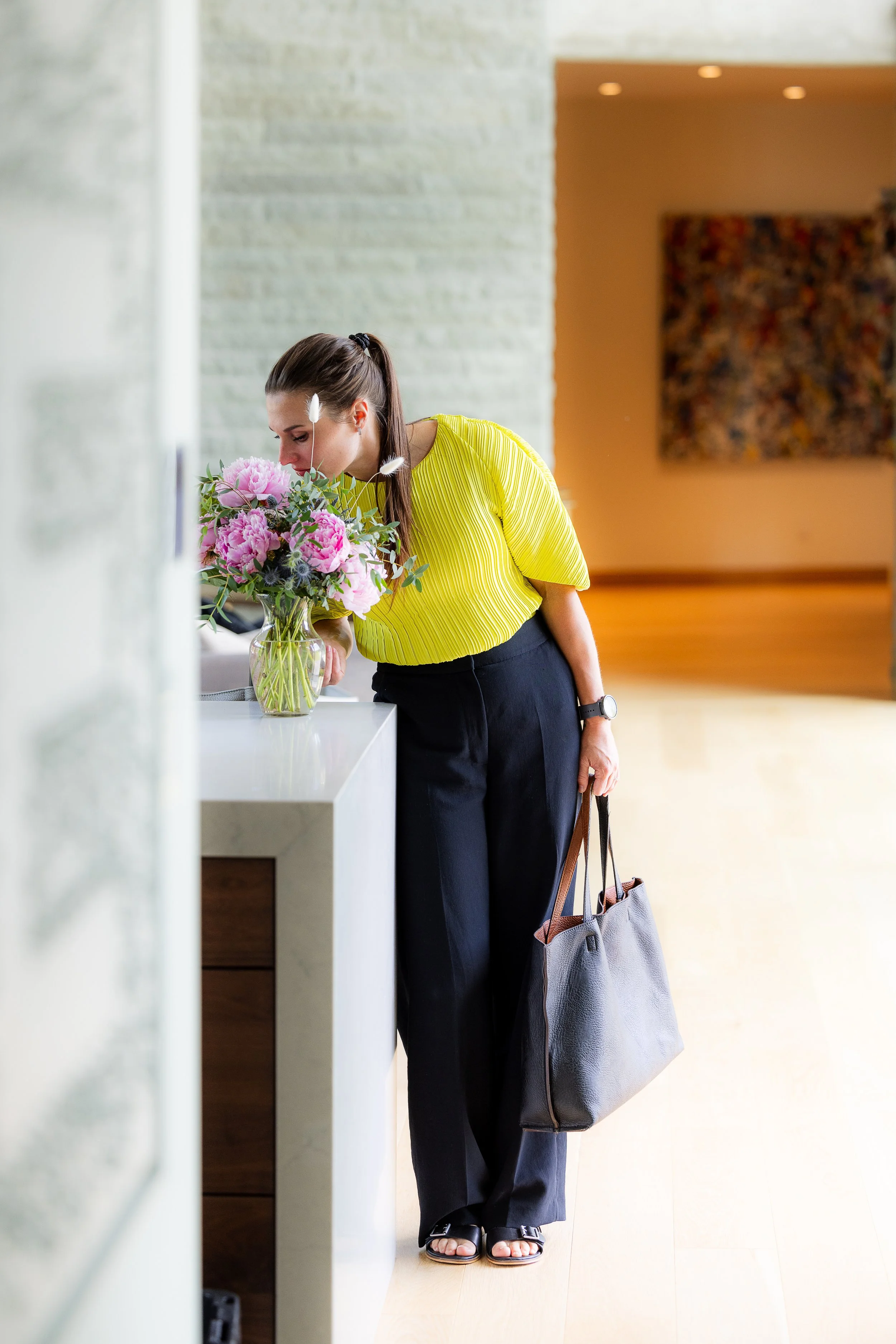 Lucy Randle standing at a desk with a bouquet of pink flowers, carrying a large gray bag and wearing sandals, in a modern indoor setting.