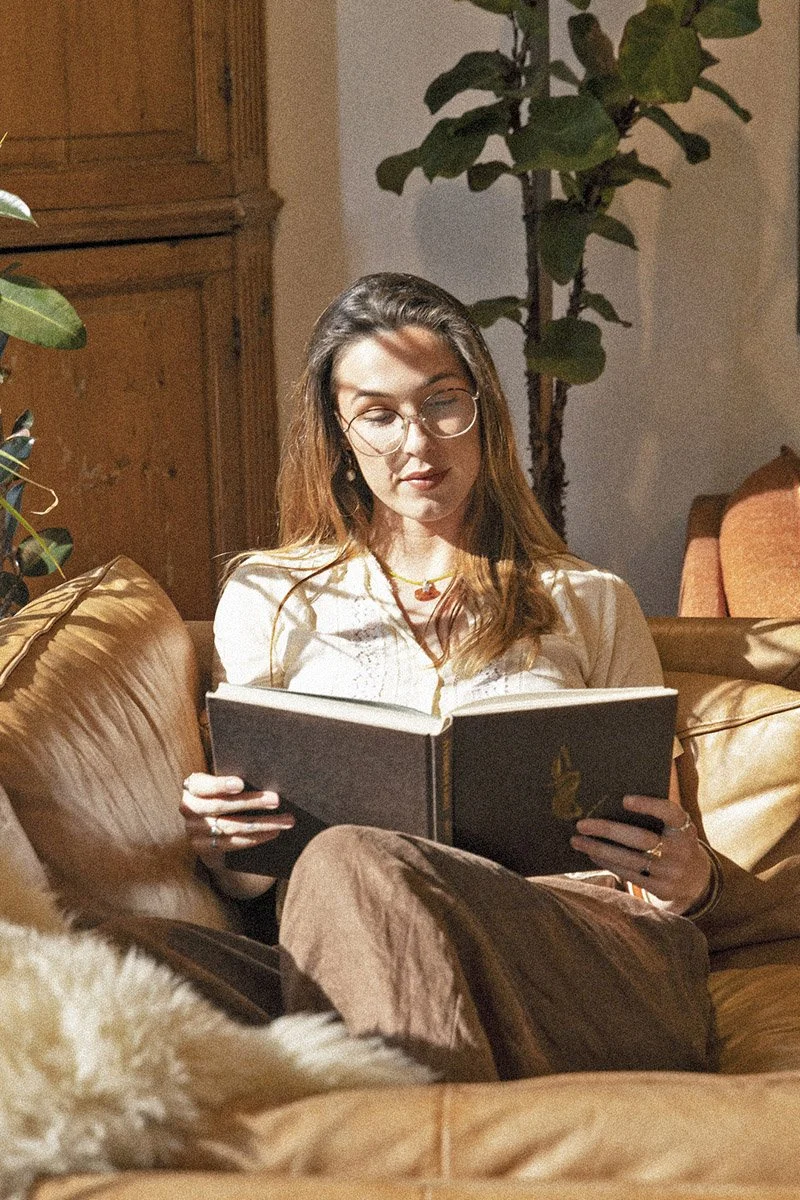 A woman with glasses and long hair sitting on a brown leather couch, reading a book in a cozy room with wooden furniture and green plants.