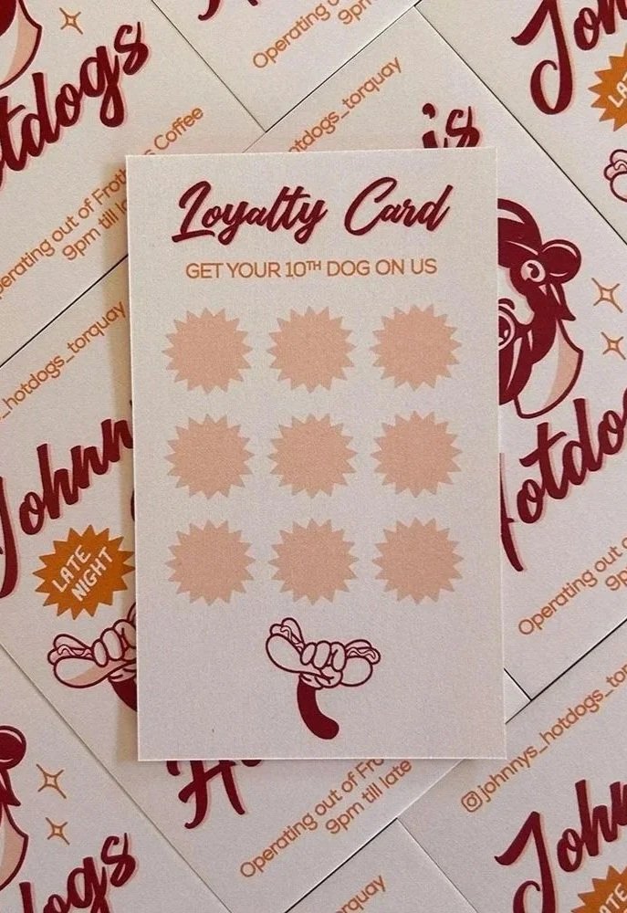 A loyalty card from Johnny's Hotdogs, with spaces for ten stamps, featuring an illustration of a hotdog at the bottom. The background shows multiple similar cards.
