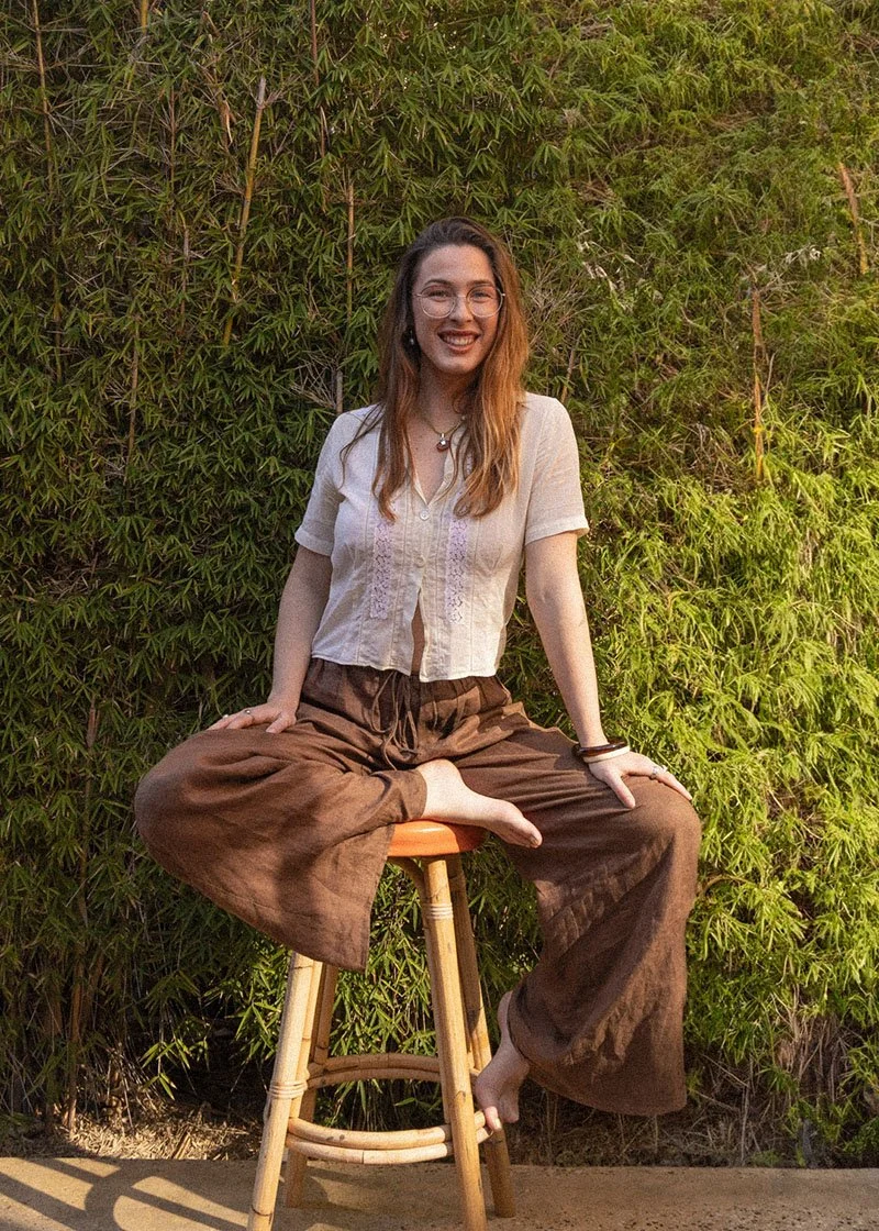A woman with long brown hair, glasses, and a necklace sits cross-legged on an orange stool in front of a lush green leafy background, smiling at the camera.
