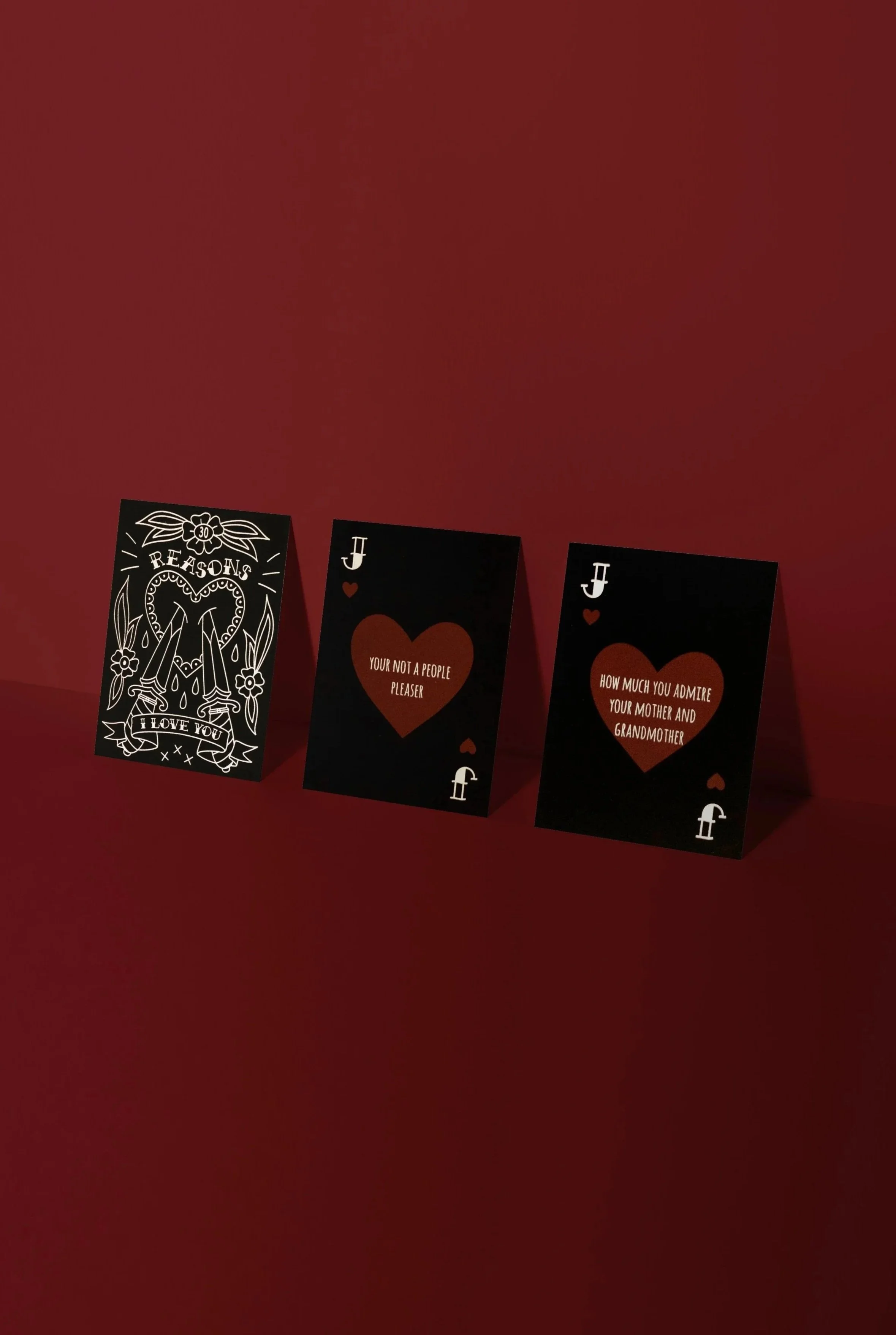 Three cards leaning against a red background that look like 2 jacks of hearts with writing on them and a cover card that says '30 Reasons I Love you'