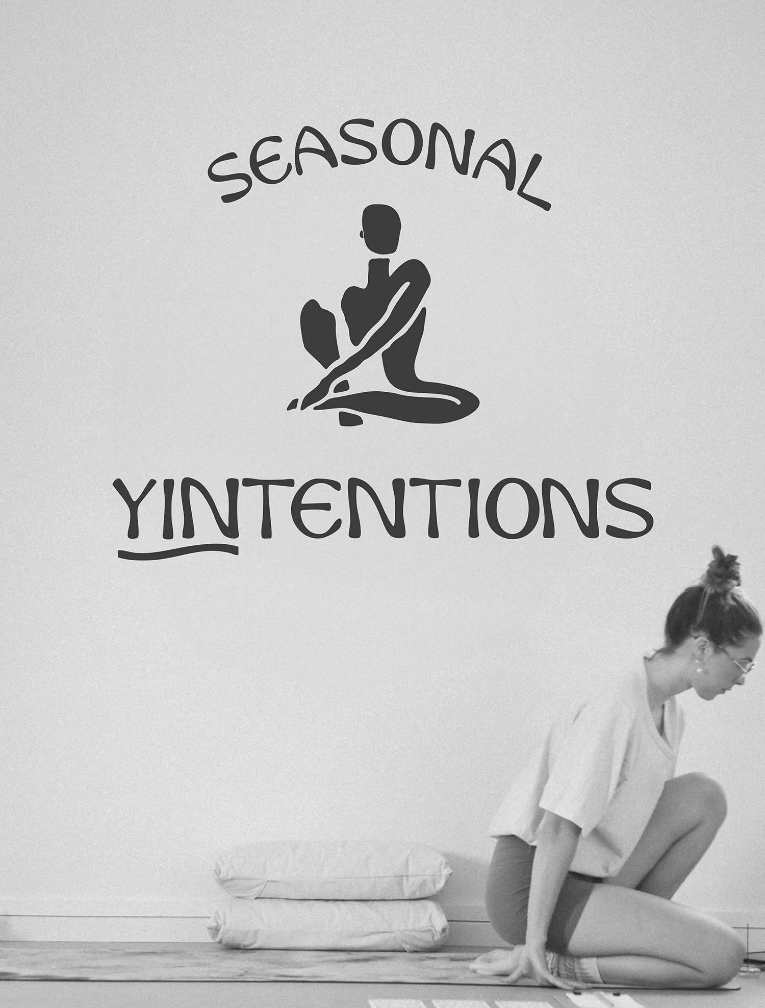 A black and white photo of a woman crouched on a yoga mat with an organic, silohette yoga logo in dark grey above her head
