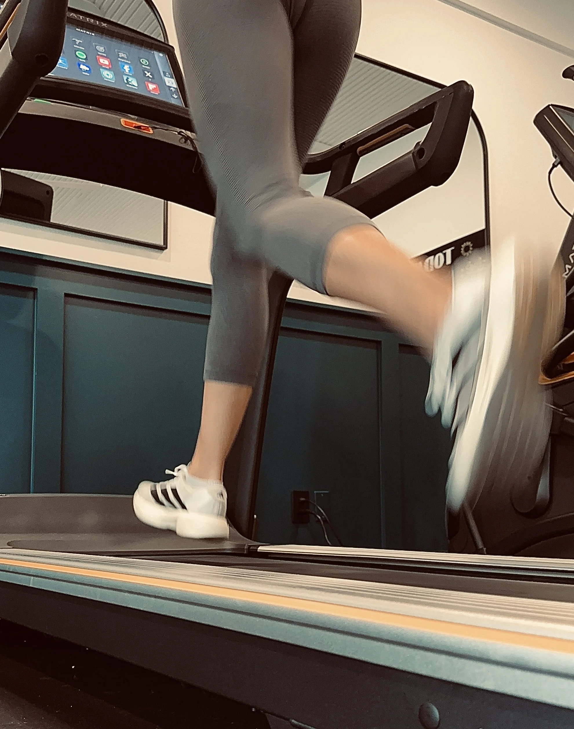 Person walking on a treadmill, wearing white sneakers with black stripes and black leggings, in a gym or fitness center.
