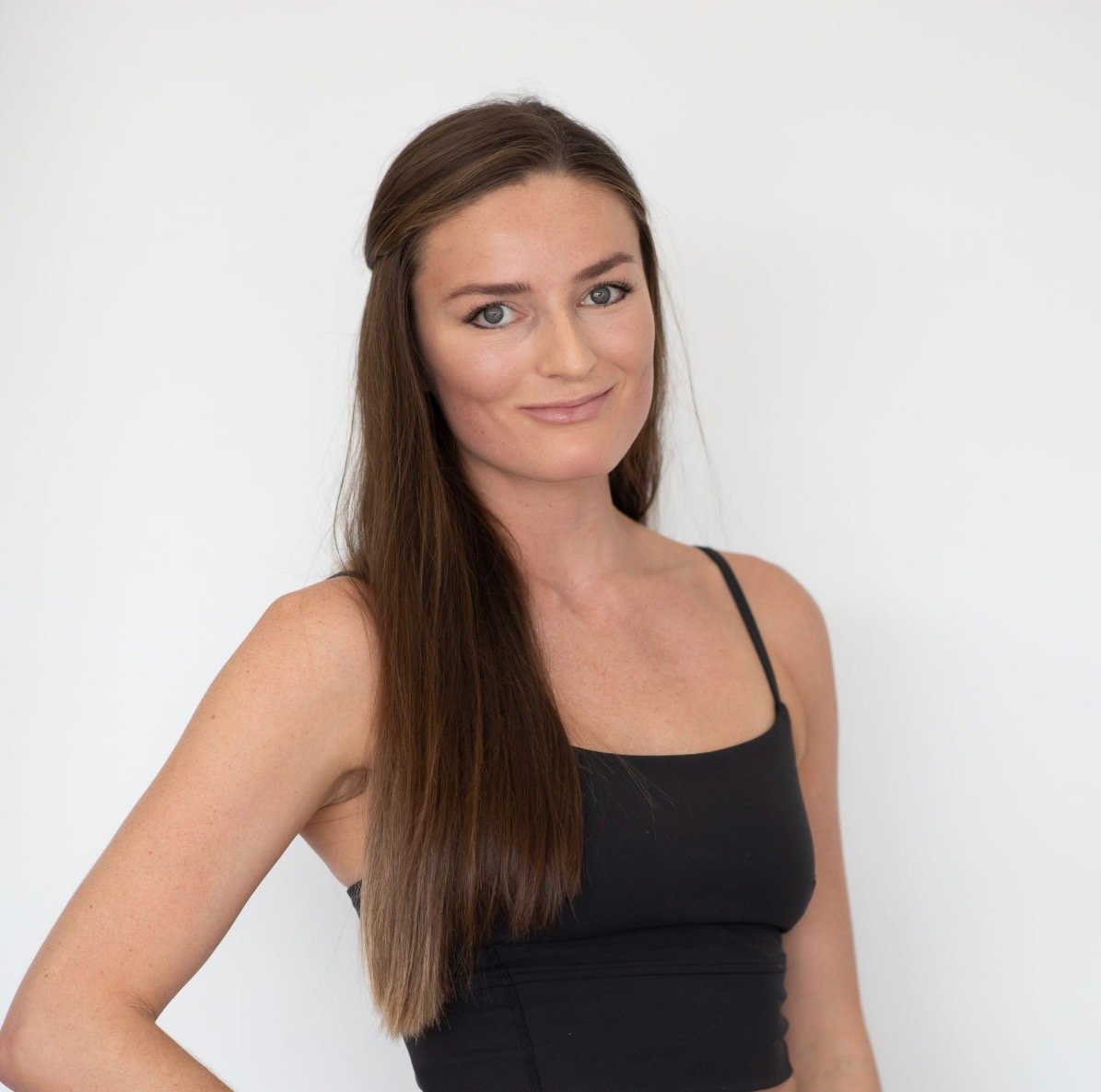 A woman with long brown hair and blue eyes wearing a black spaghetti strap top, standing against a plain white background, smiling softly.