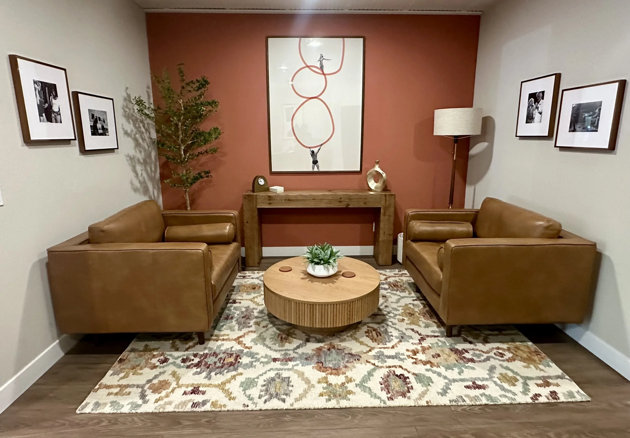 A living room with two leather sofas facing each other, a wooden coffee table with a plant on top, a patterned rug, a large framed artwork of a person balancing on red rings on the wall, a green plant on the left, a tall floor lamp on the right, and framed photographs hanging on the beige and reddish-brown walls.