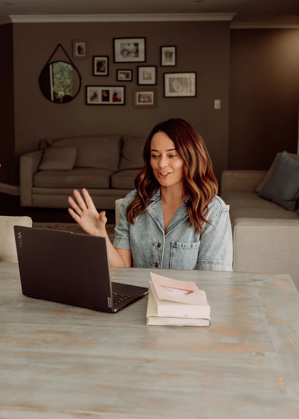 A woman with wavy brown hair in a denim shirt is sitting at a table, waving while talking on a video call on her laptop. There are books on the table, and a cozy living room with framed photos on the wall behind her.