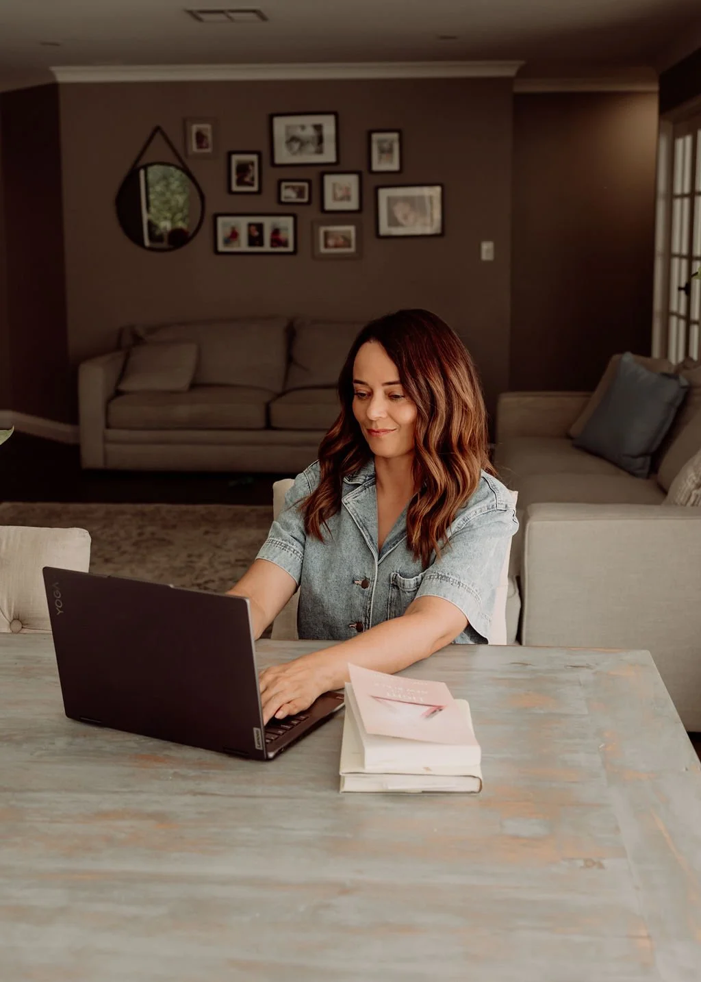 A woman with shoulder-length brown hair, wearing a light blue denim shirt, is sitting at a wooden table working on a black Lenovo Yoga laptop. There is a closed book and a notebook on the table in front of her. The background features a gray living room with a beige sofa, framed family photos on the wall, and a mirror.