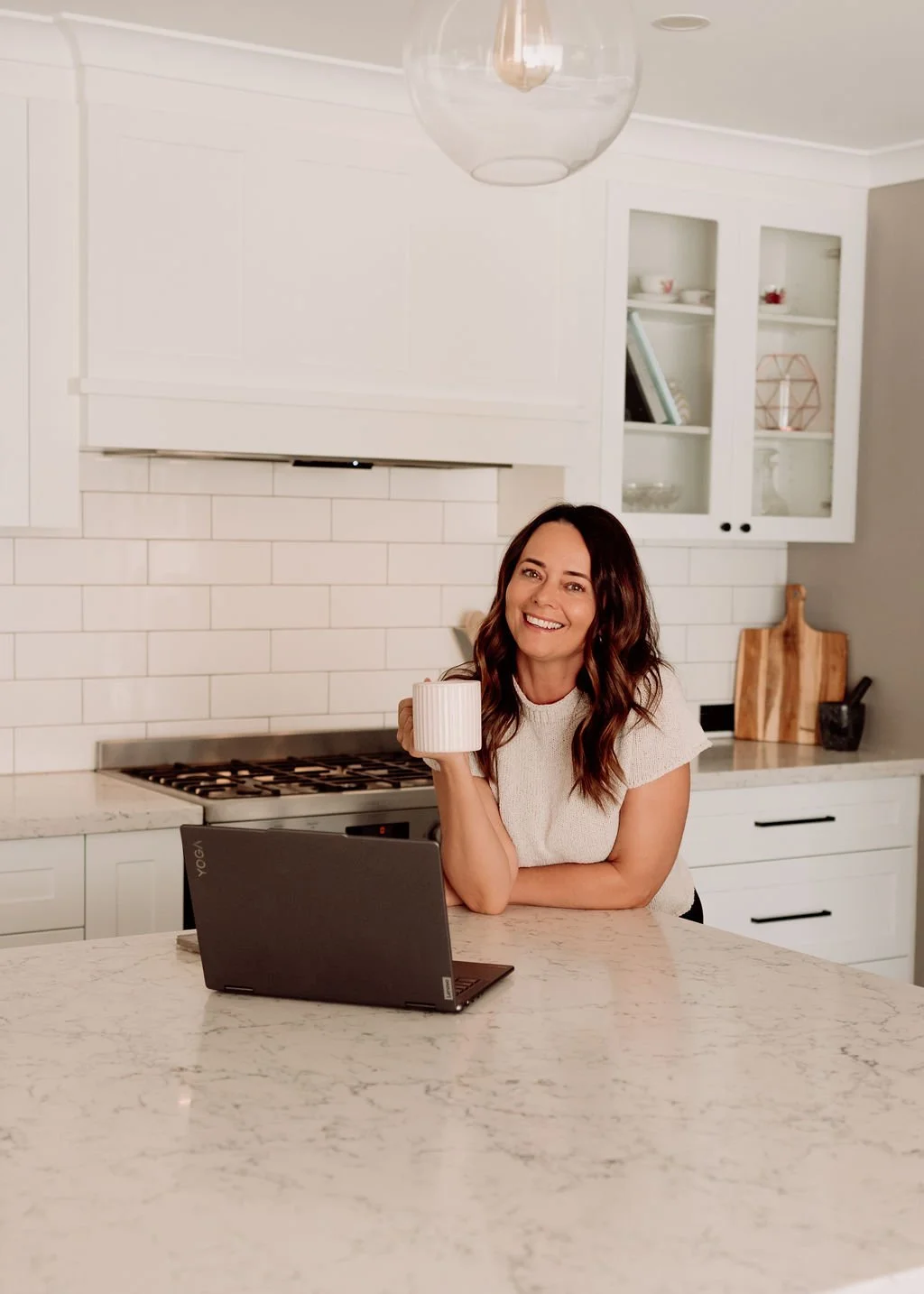 A woman smiling and holding a mug in a modern kitchen, sitting in front of a laptop on a marble countertop.