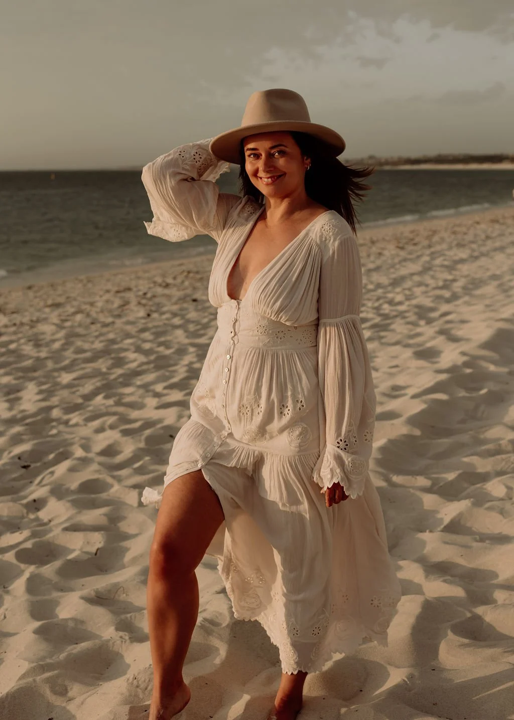 A woman standing barefoot on a sandy beach, smiling, wearing a white dress with embroidery and a wide-brimmed hat, with the ocean and cloudy sky in the background.