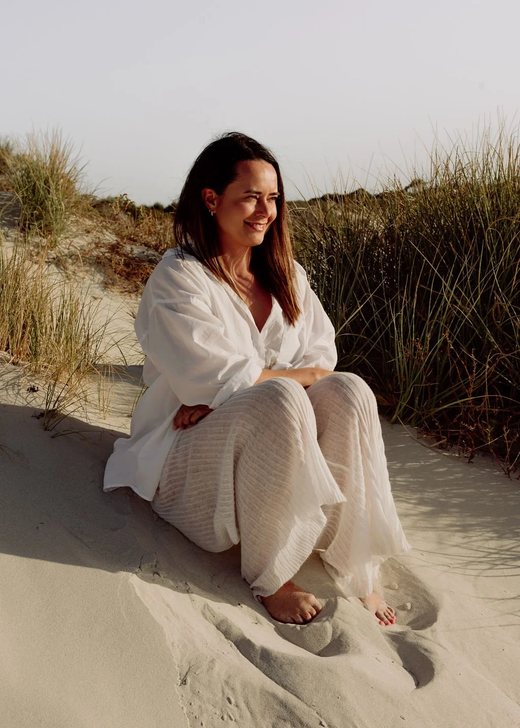 A woman sitting on sandy dunes near beach grass, smiling and enjoying the outdoors.