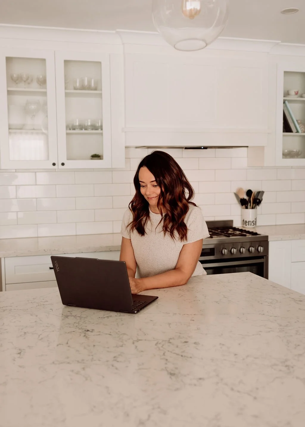 A woman sitting at a kitchen island using a laptop, with white cabinets and a stove in the background.