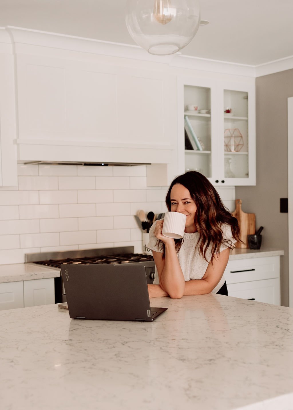 Woman drinking coffee in a modern kitchen while using a laptop.