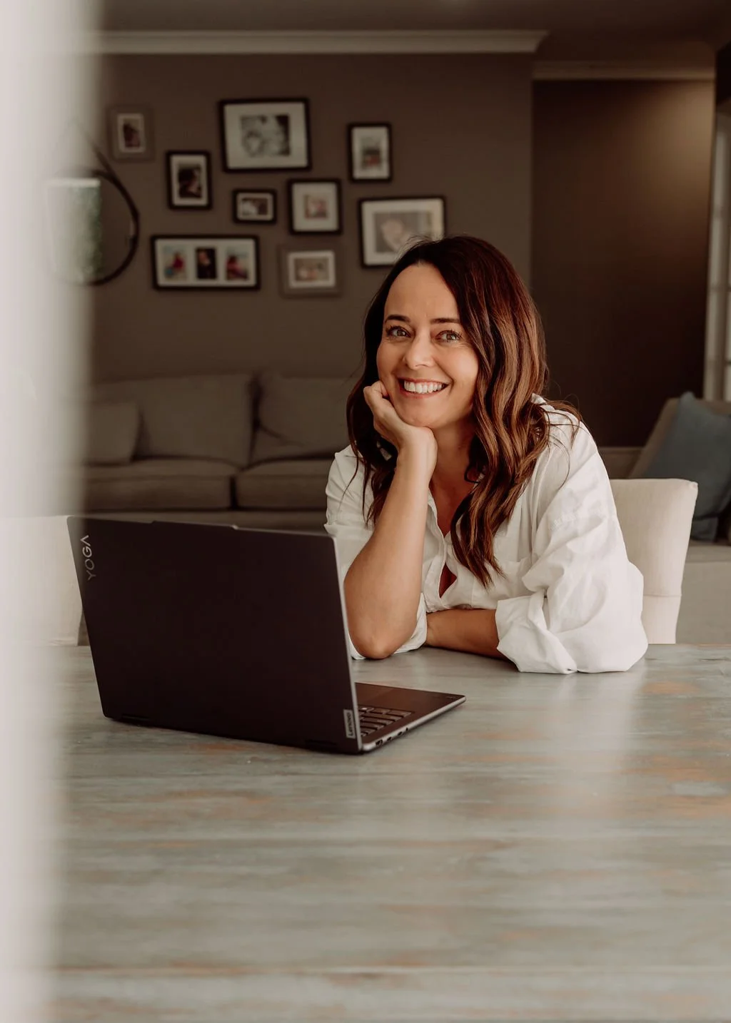 A woman with long brown hair smiling while sitting at a table with a laptop, in a living room with a gallery wall of picture frames in the background.