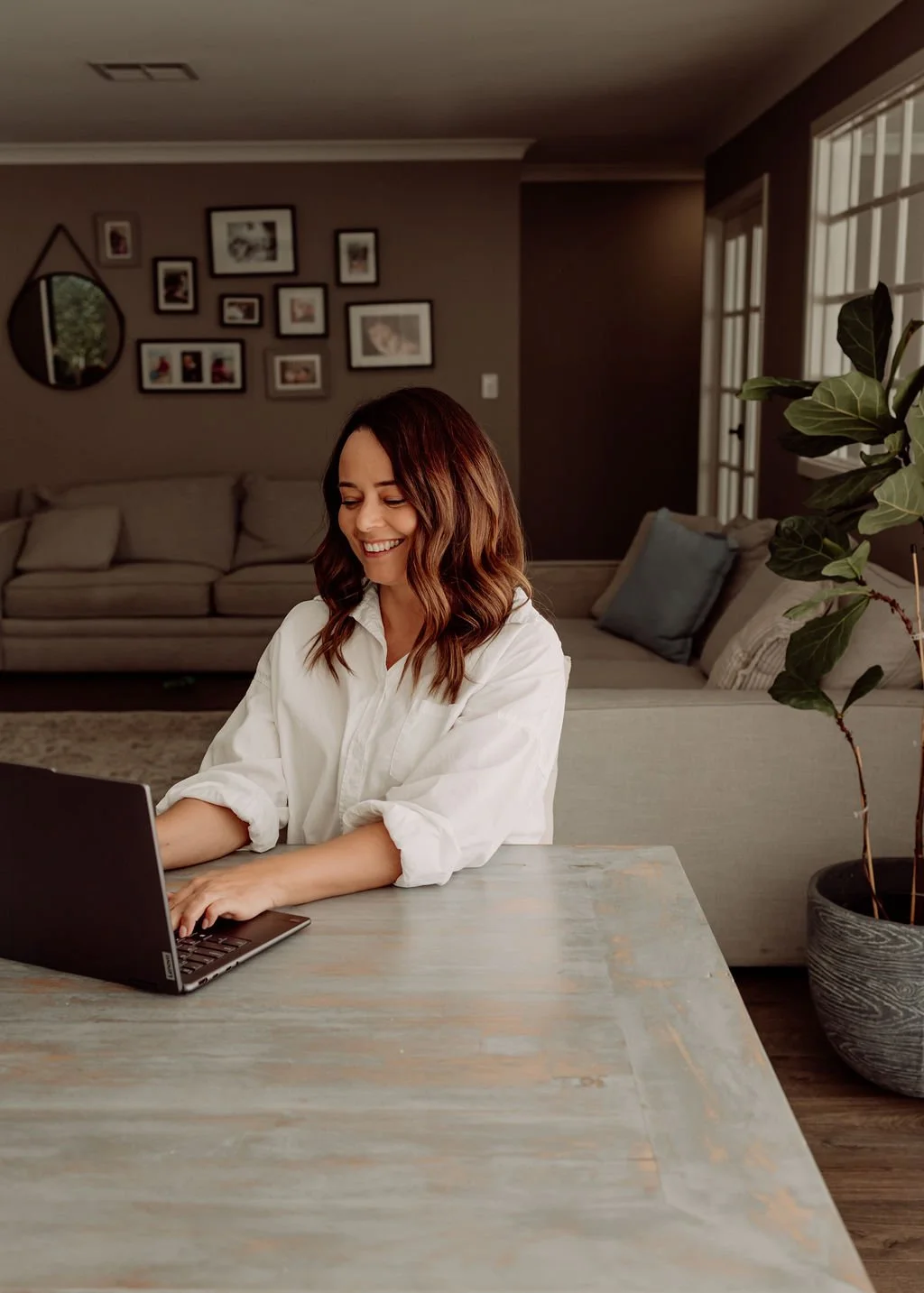 A woman with brown hair smiling while working on a laptop at a dining table in a cozy living room.