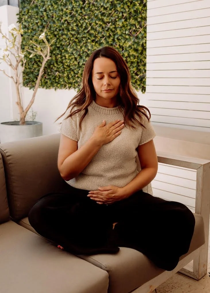 A woman sitting cross-legged on a couch with her eyes closed, one hand on her chest and the other on her abdomen, practicing mindfulness or meditation in a cozy room with a green plant and white wooden wall in the background.