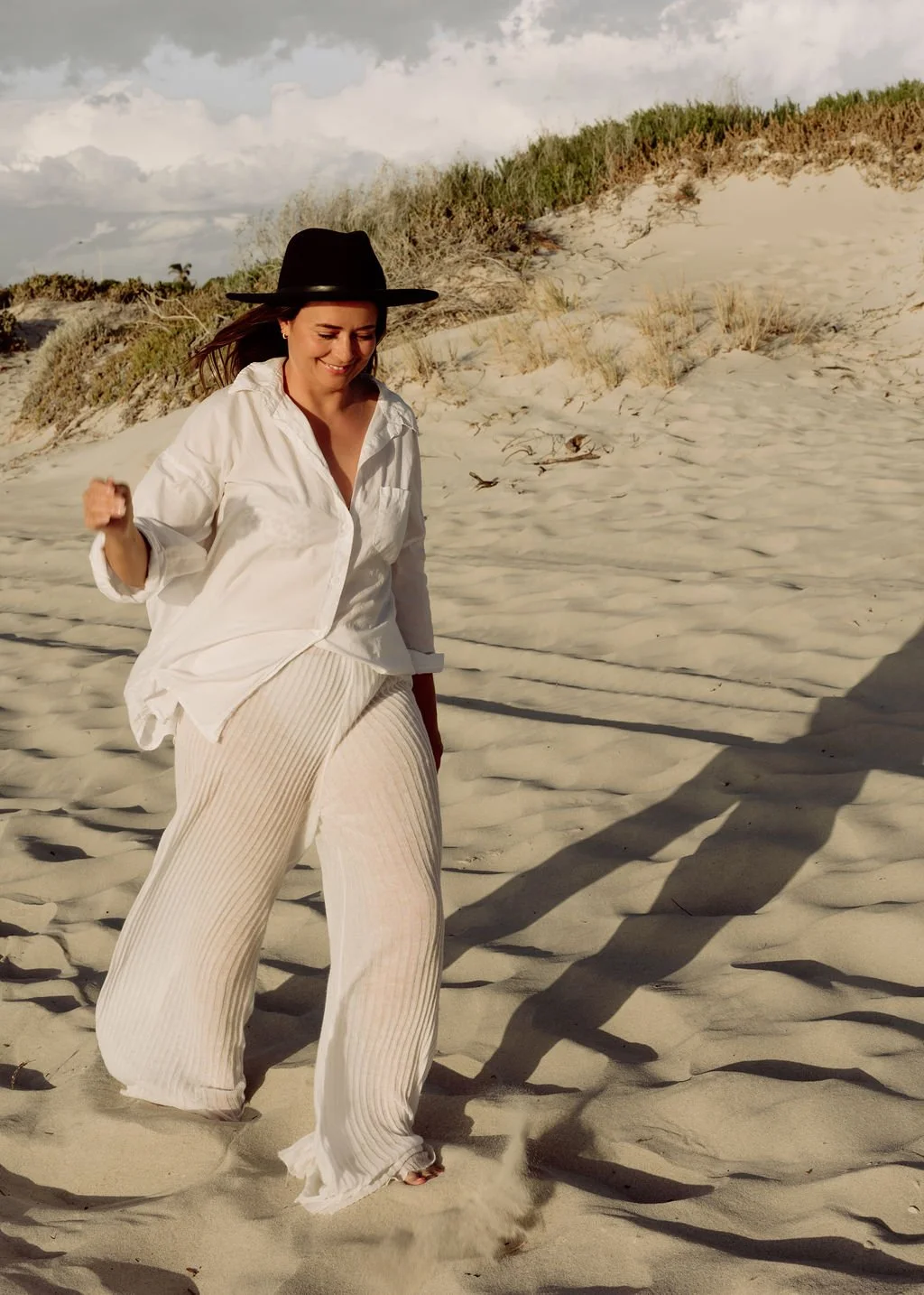 A woman in a white shirt, cream-colored pants, and a black hat walking on a sandy beach with a smile, casting a long shadow.