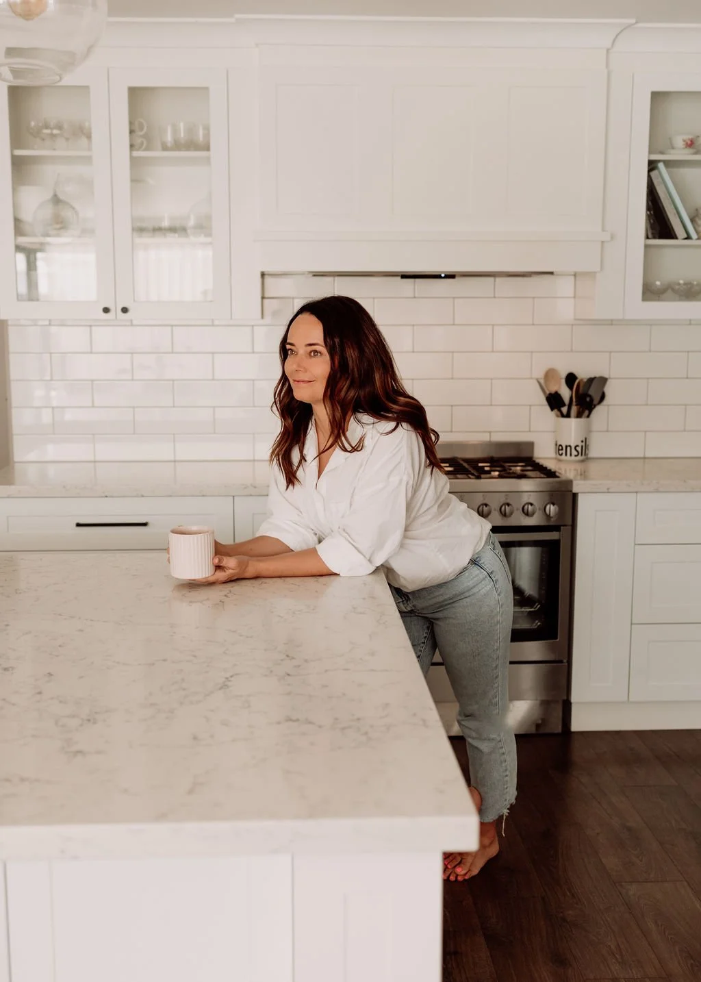 A woman with long brown hair wearing a white shirt and gray jeans leaning on a kitchen counter with a cup in her hand, in a modern white kitchen with white cabinets, white subway tile backsplash, and a stainless steel stove.