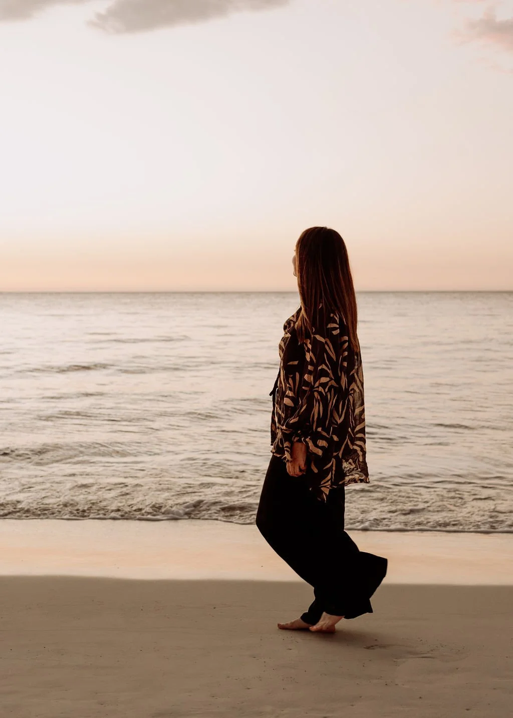 A woman standing barefoot on the beach during sunset, looking at the ocean.