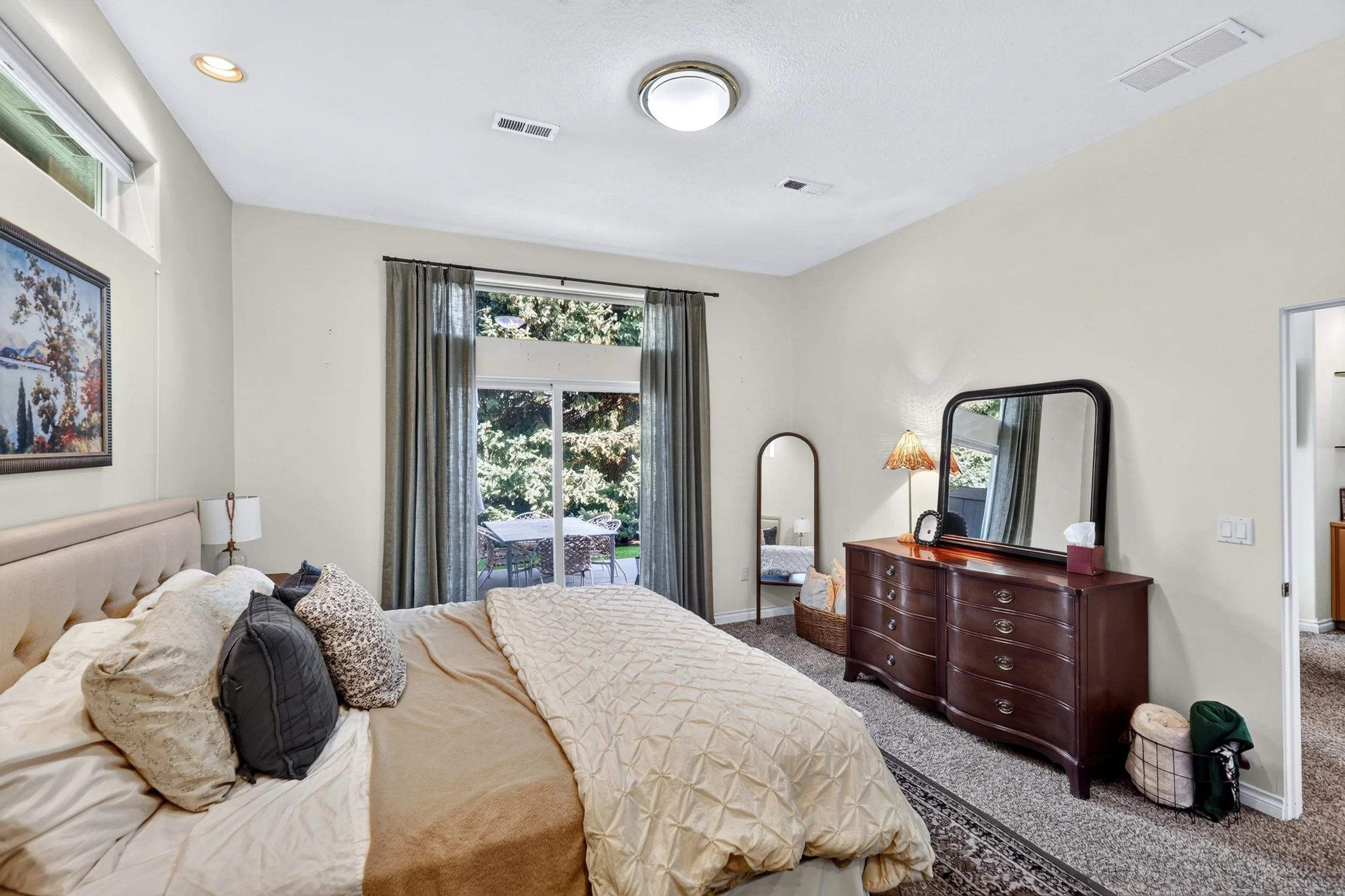 A bedroom with a large bed, cream-colored bedding, and multiple pillows, a wooden dresser with a mirror, and a sliding glass door with curtains leading to an outdoor patio with a table and chairs, surrounded by greenery.