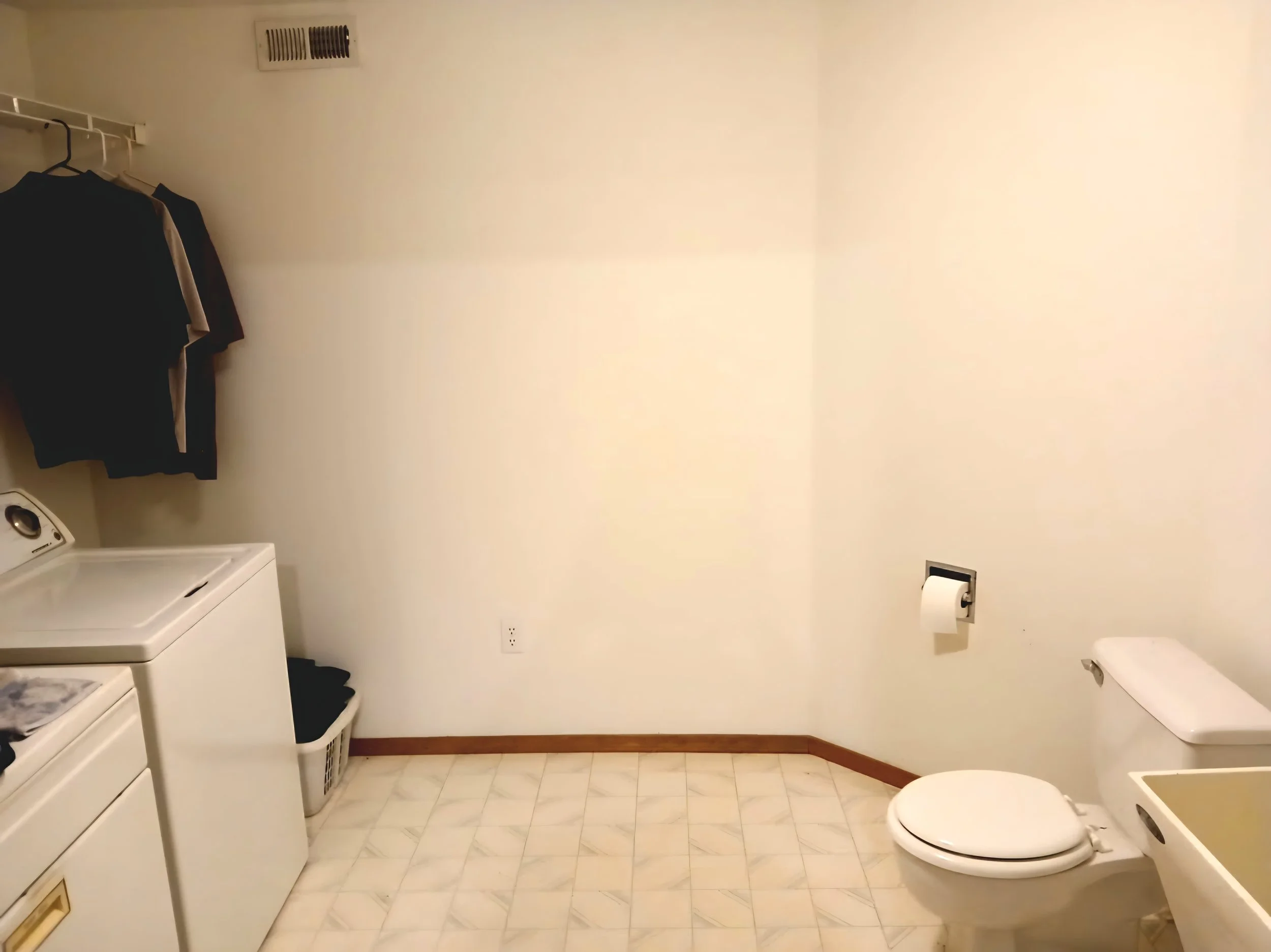 A bathroom with a washing machine, dryer, and laundry basket on a tiled floor, a towel hanging on a rack, a toilet paper holder with a roll, and a toilet.