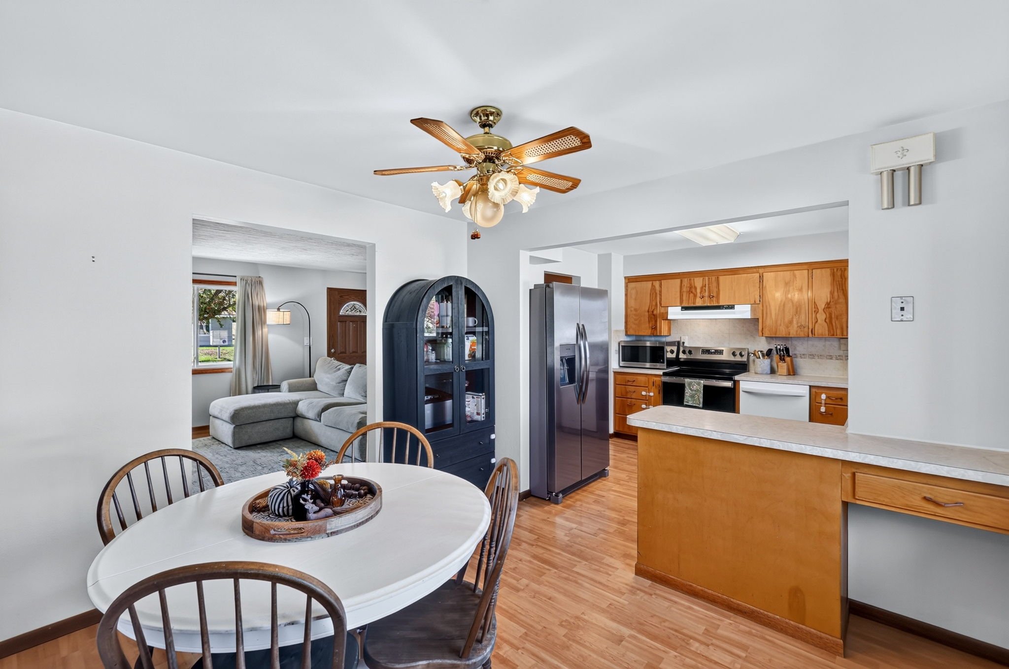 View of a living room and kitchen with white walls and wooden flooring, a ceiling fan with wooden blades, a round dining table with chairs, a black display cabinet, stainless steel refrigerator, microwave, and wooden cabinets in the kitchen.