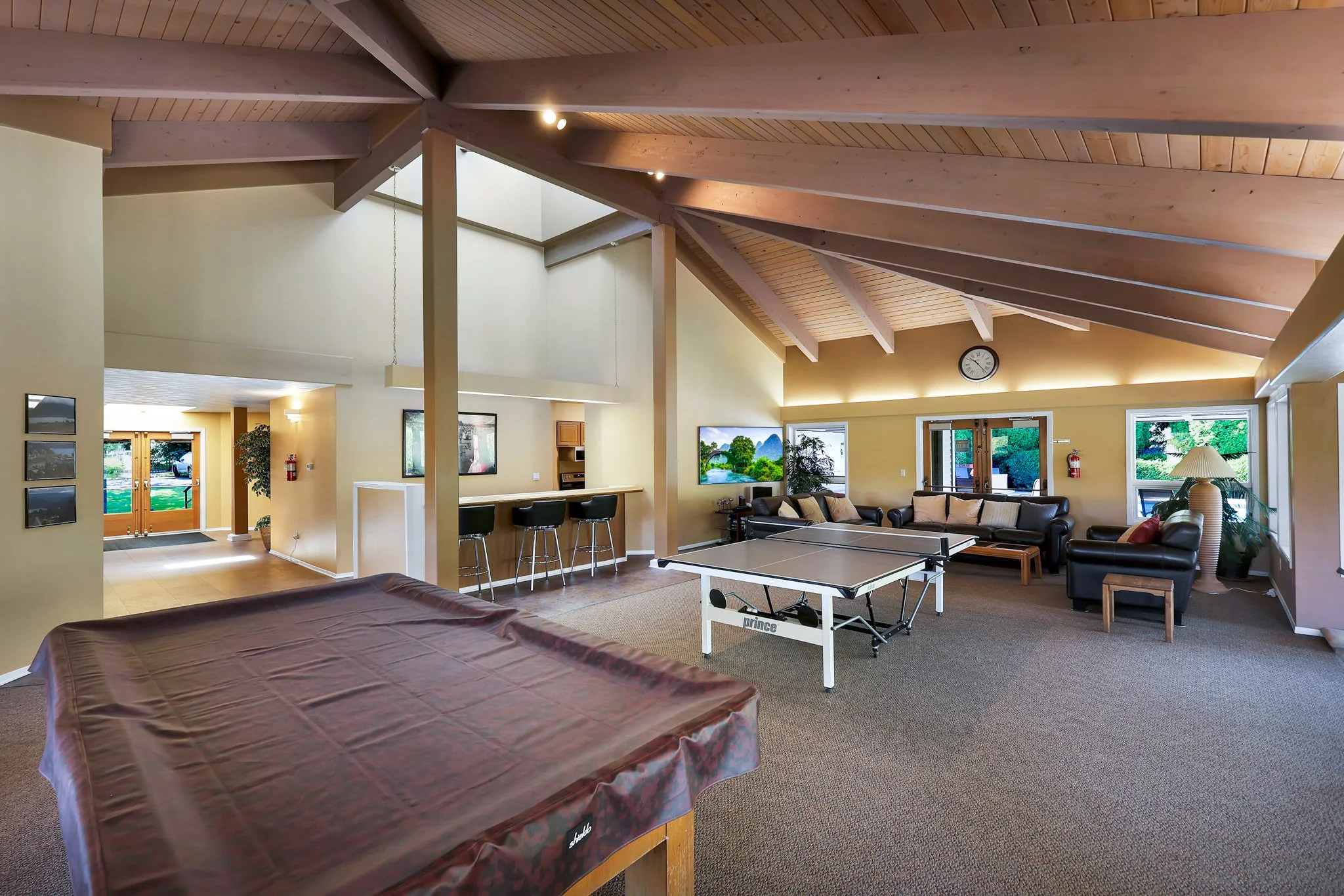 Interior of a spacious living area with a high wooden ceiling, multiple windows, a clock on the wall, and various furniture including a ping pong table, sofas, and a table with a brown cover.