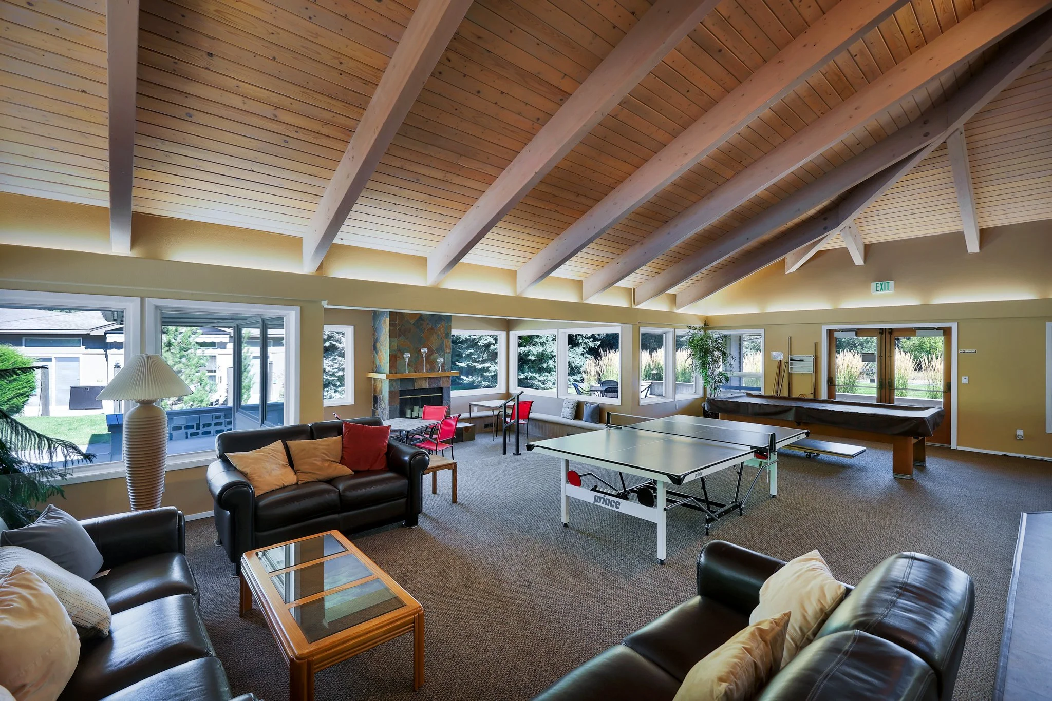 Living room with large windows, a fireplace, several sofas, a coffee table, and a ping pong table, with a wooden vaulted ceiling.