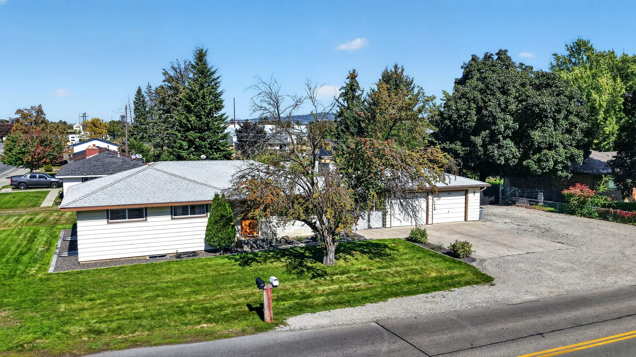 A suburban house with a white exterior, gray roof, and a detached garage. A large tree with sparse leaves stands in the front yard, surrounded by a well-maintained green lawn. There is a driveway leading to the garage and a sidewalk along the street.