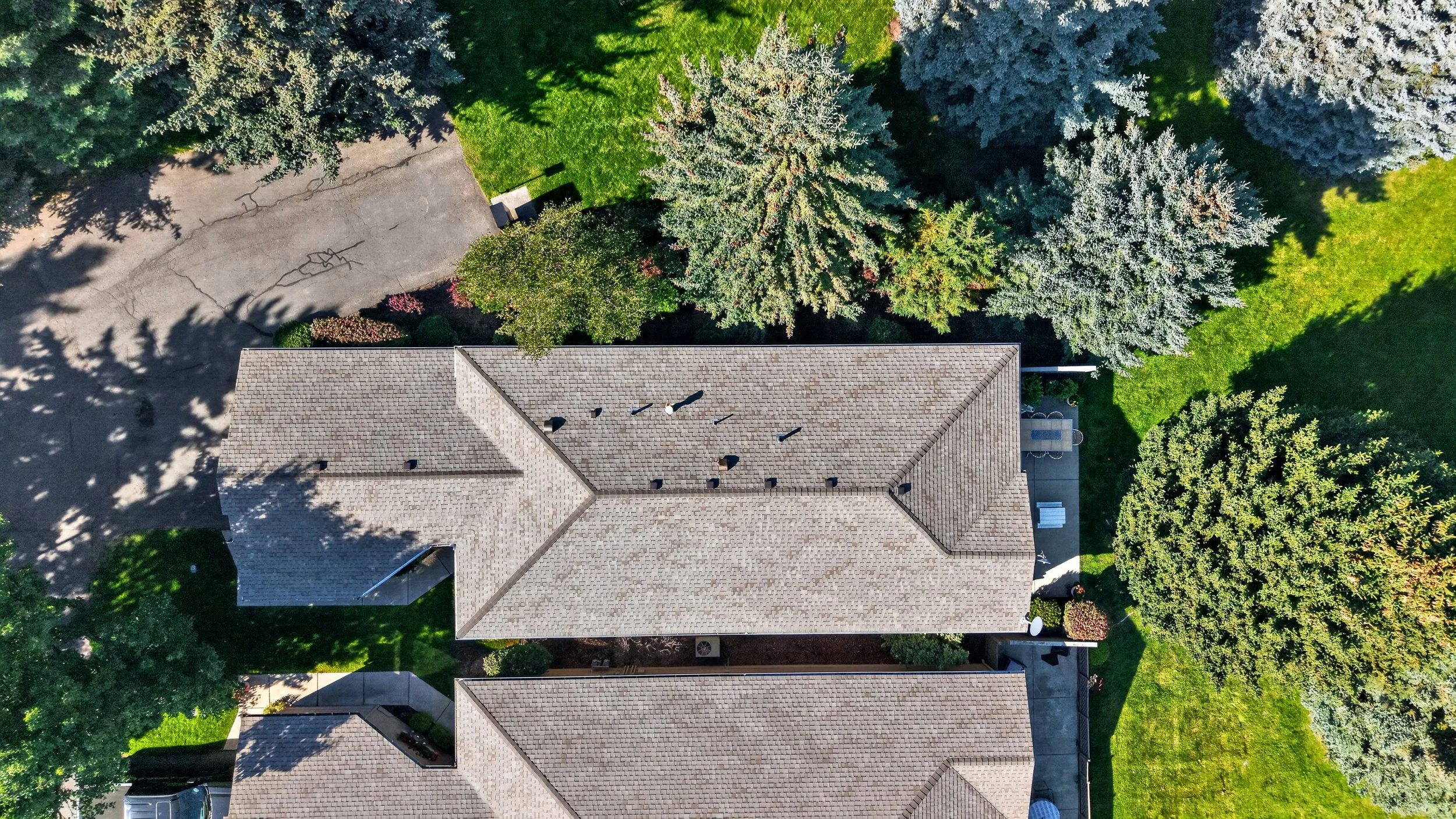 An aerial view of a house surrounded by green trees and a grassy yard. The house has a shingled roof with multiple vents, and a concrete driveway leading to the side. There are colorful bushes near the edge of the house.