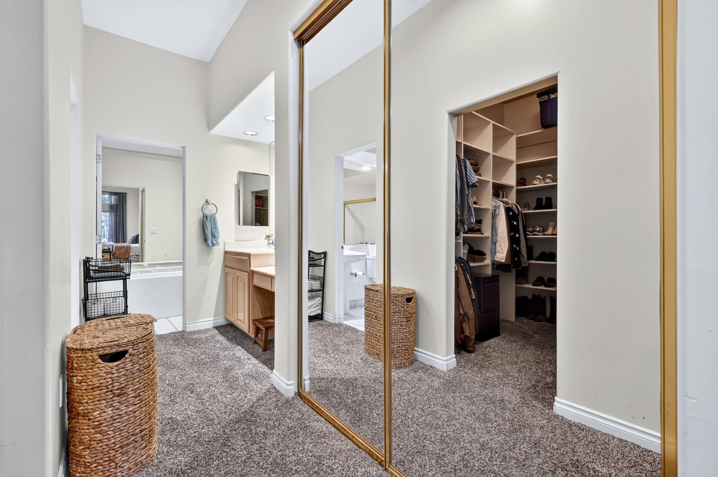View of a walk-in closet with a mirrored door, open shelves with shoes, hanging clothes, and a laundry hamper. A partial bathroom is visible through the doorway.