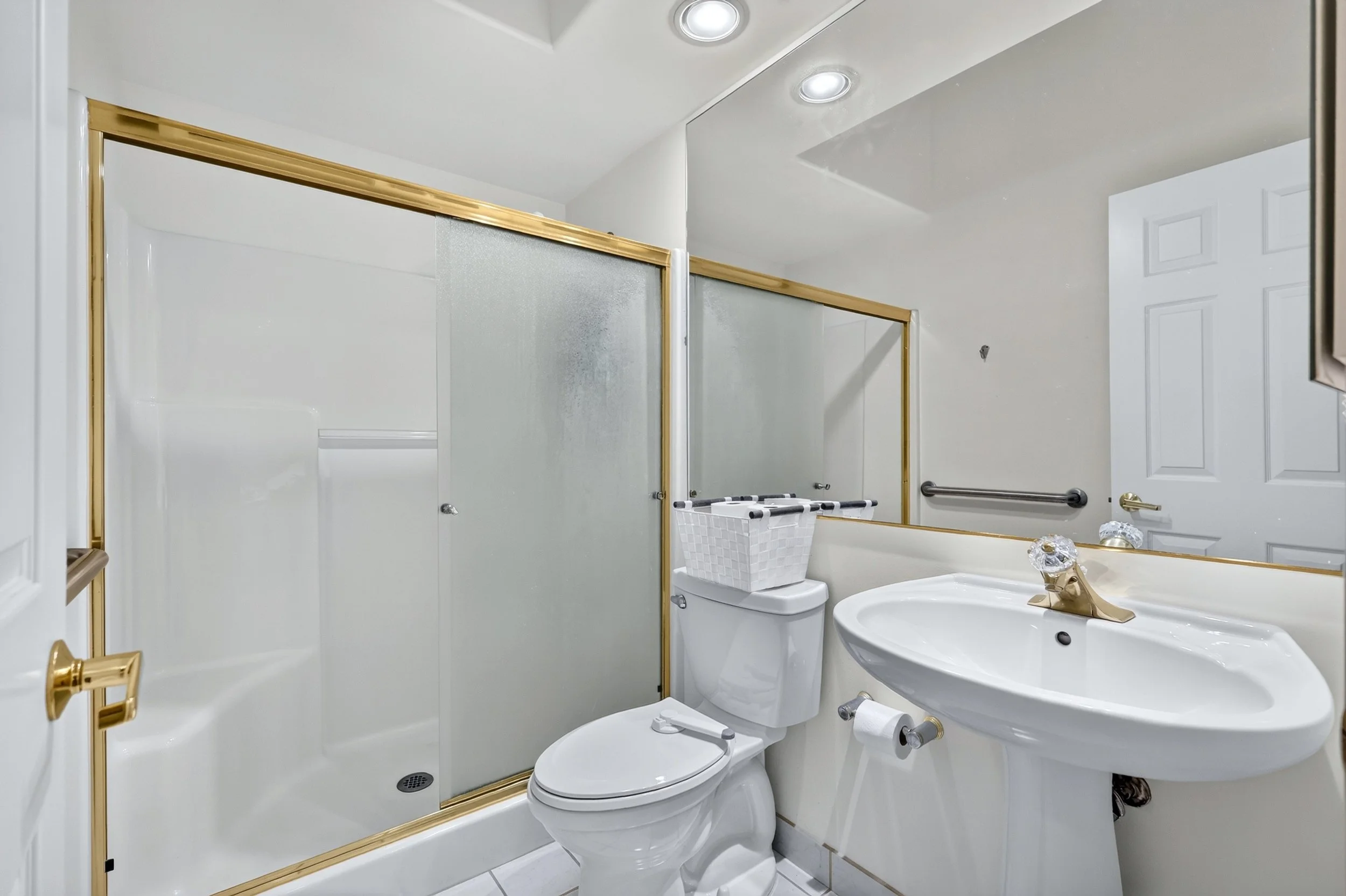 A small bathroom featuring a shower with frosted glass doors and gold trim, a white toilet with a laundry basket on top, and a white sink with gold fixtures. The walls are white, with a towel bar and a mirror above the sink, and the ceiling has reces