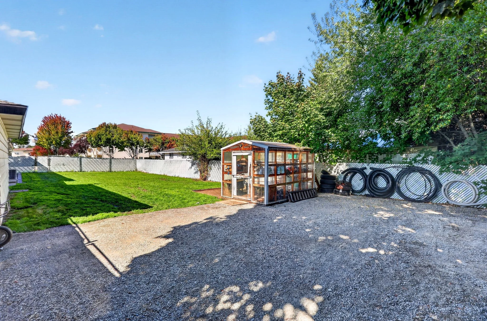 A backyard with a green lawn, a white fence, trees with fall foliage, and a small wooden shed. There are black tires and white tubing stacked on the gravel ground, with a house and more trees in the background under a clear blue sky.