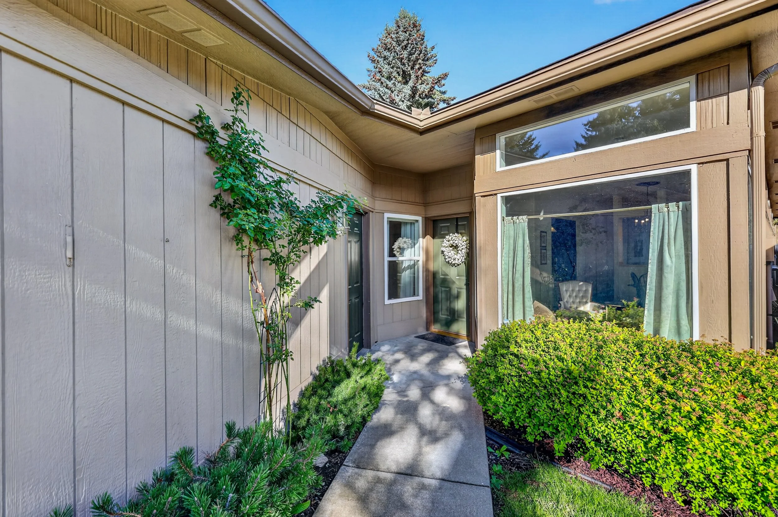 Front porch of a house with beige and brown siding, large window with curtains, a door decorated with a wreath, and a concrete pathway surrounded by green bushes and plants, with a tall tree and blue sky in the background.