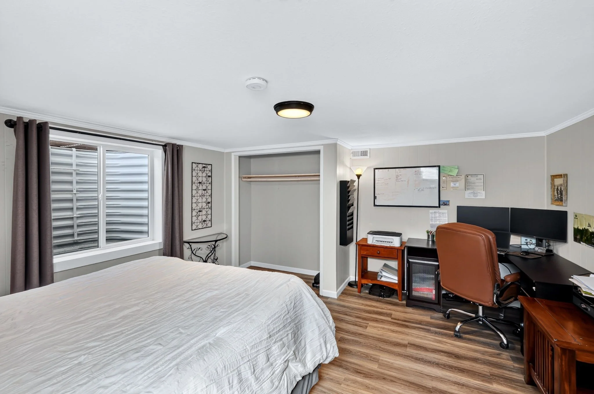 A bedroom with a bed, window with brown curtains, and an office area with a computer, whiteboard, and office furniture.