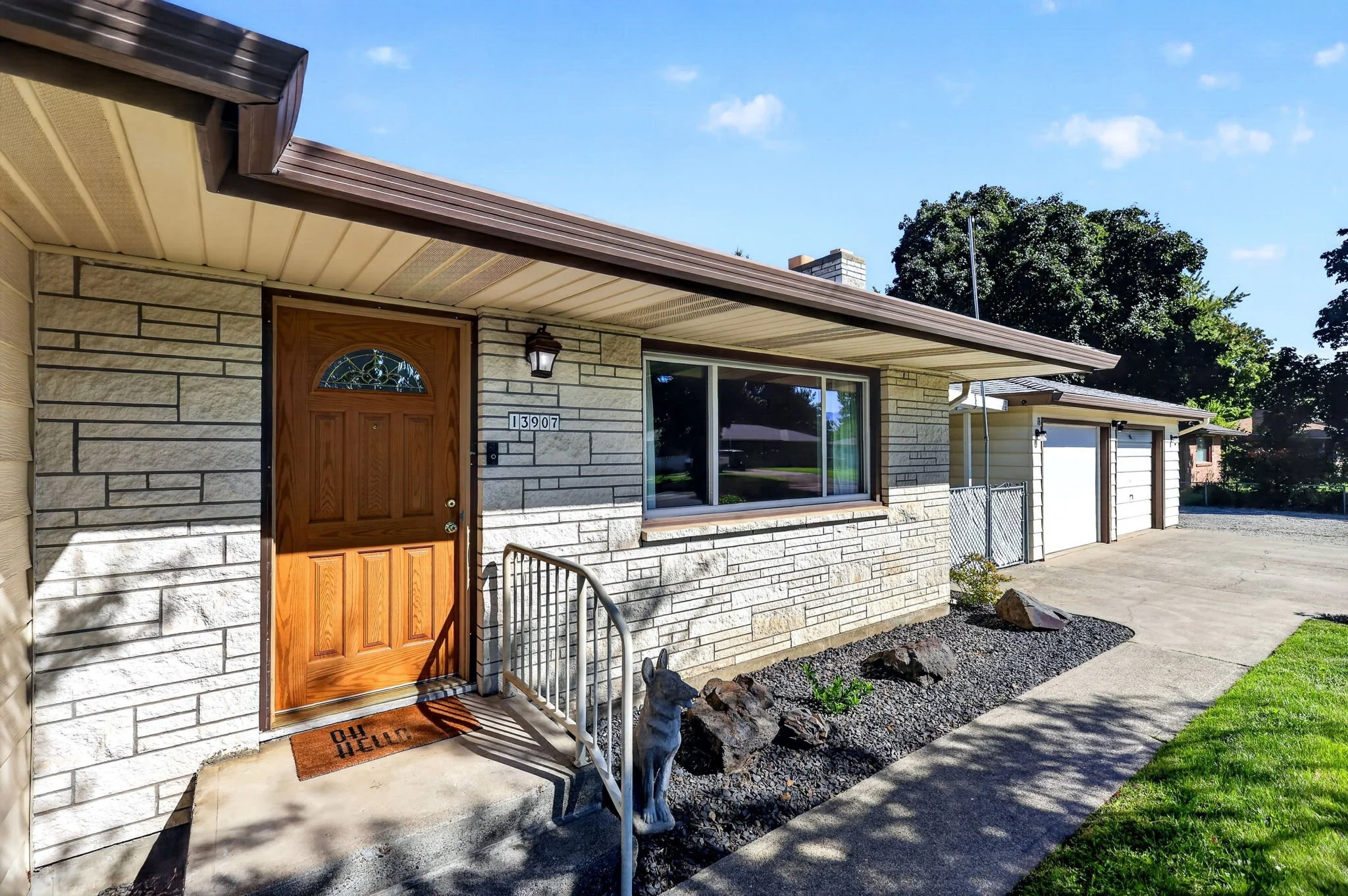 Front exterior of a house with a brick facade, wooden front door with decorative glass, a security camera, house number 13907, a porch light, and a doormat that reads 'HELLO'. There is a large window next to the door, a small stone sculpture of a dog