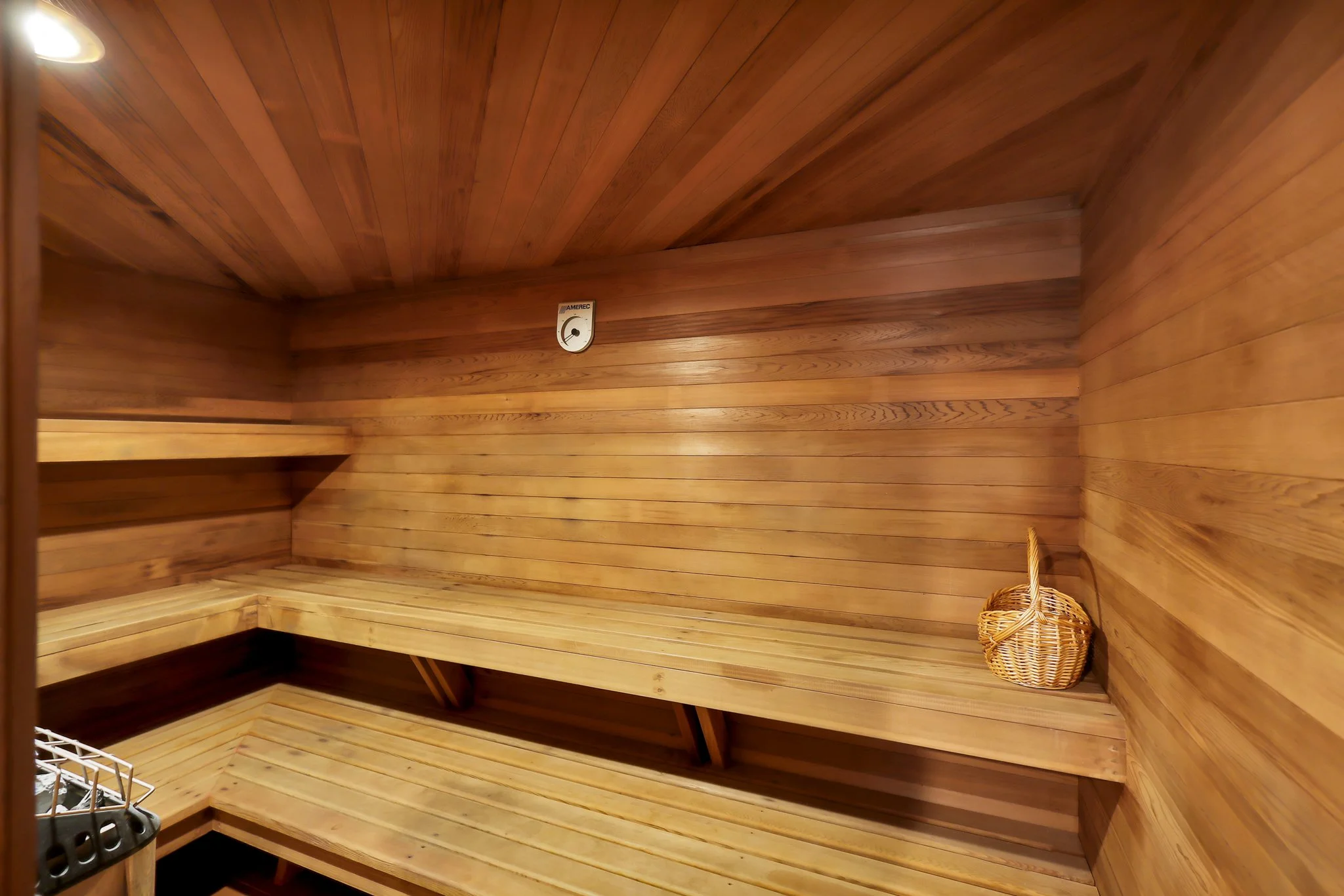 Wood-paneled interior of a sauna room with a small wicker basket in the corner and a metal heater at the bottom left.