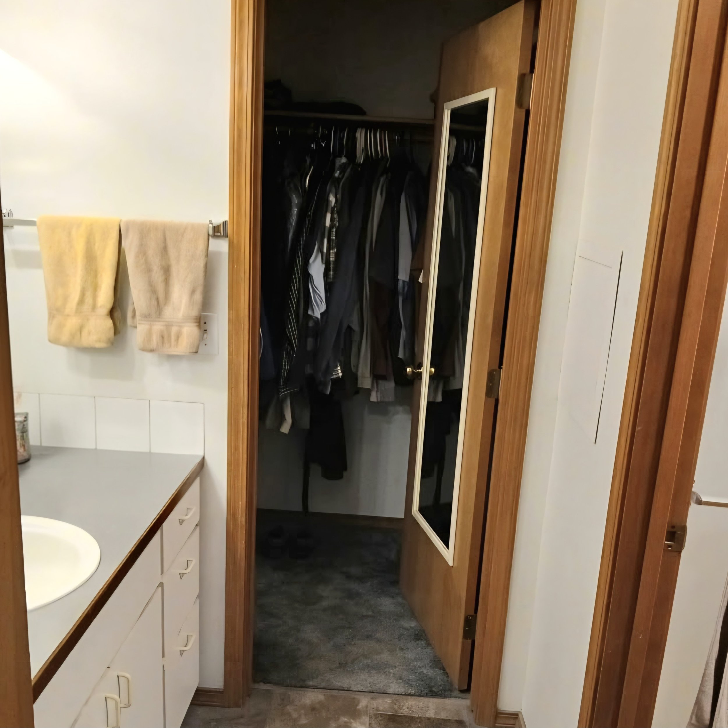 A closet with hanging clothes and a mirror on the door, part of a room with wooden trim and a countertop with towels.