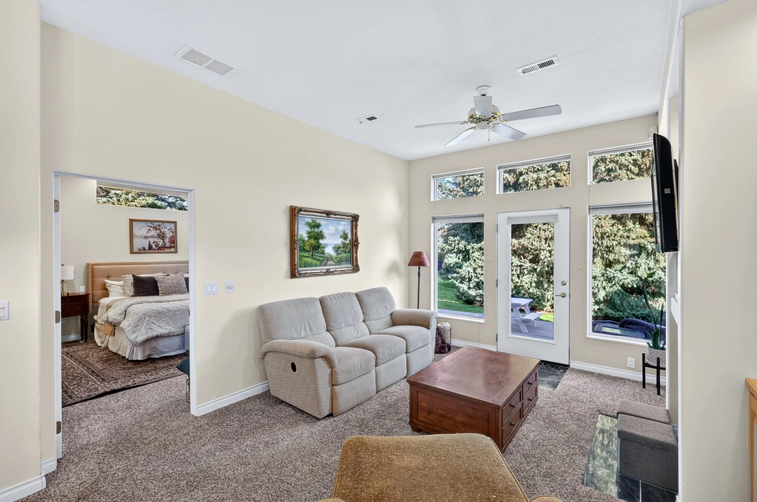 Living room with beige sofa, wooden coffee table, large windows showing trees, and a view into a bedroom with a bed and framed artwork. There is a ceiling fan and a television mounted on the wall, with a door leading outside to a patio with outdoor c