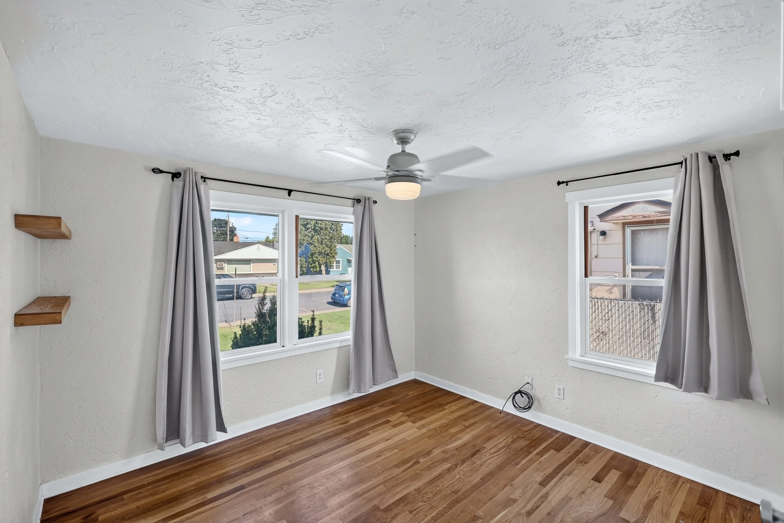Empty room with white walls, wooden floor, two large windows with white curtains, ceiling fan with light, and small black shelf on the left wall.