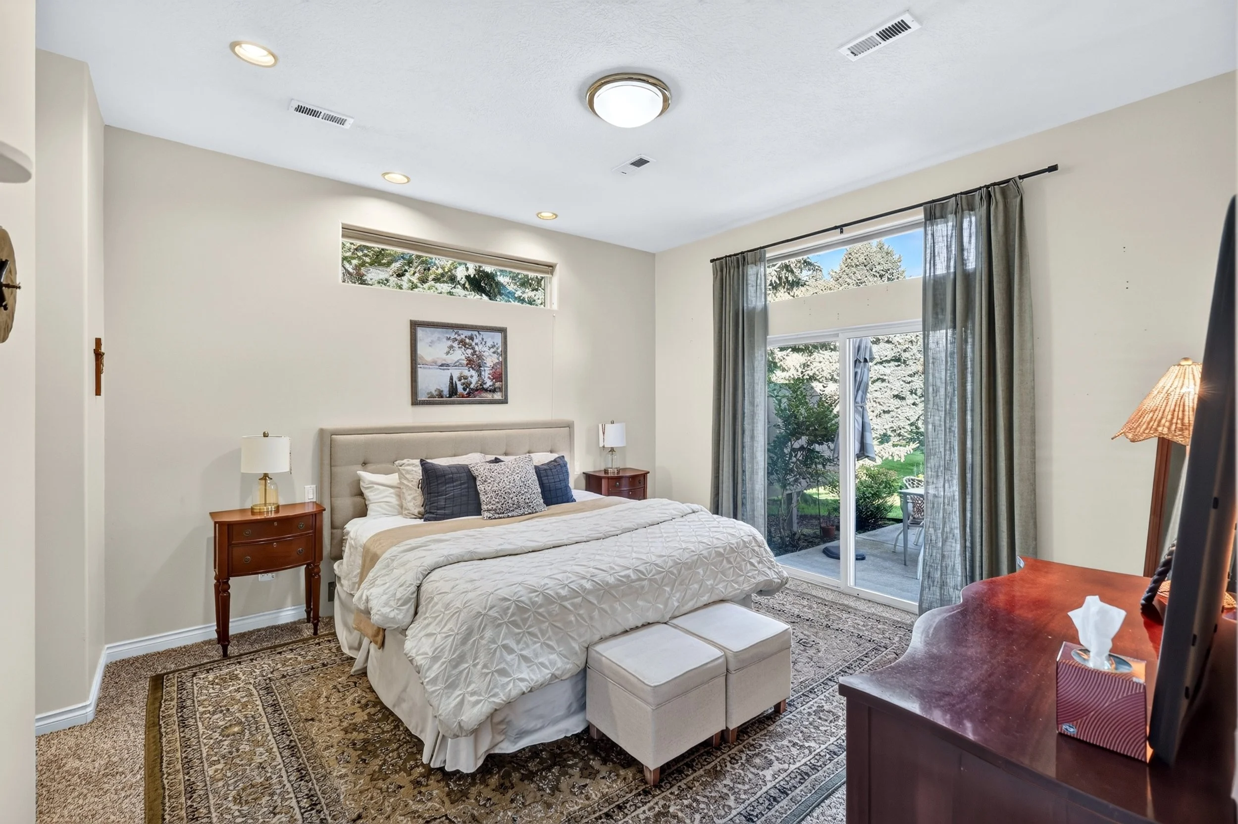 A bedroom with a bed, side tables, lamps, a painting, windows with curtains, and a sliding glass door leading to a patio with outdoor furniture.