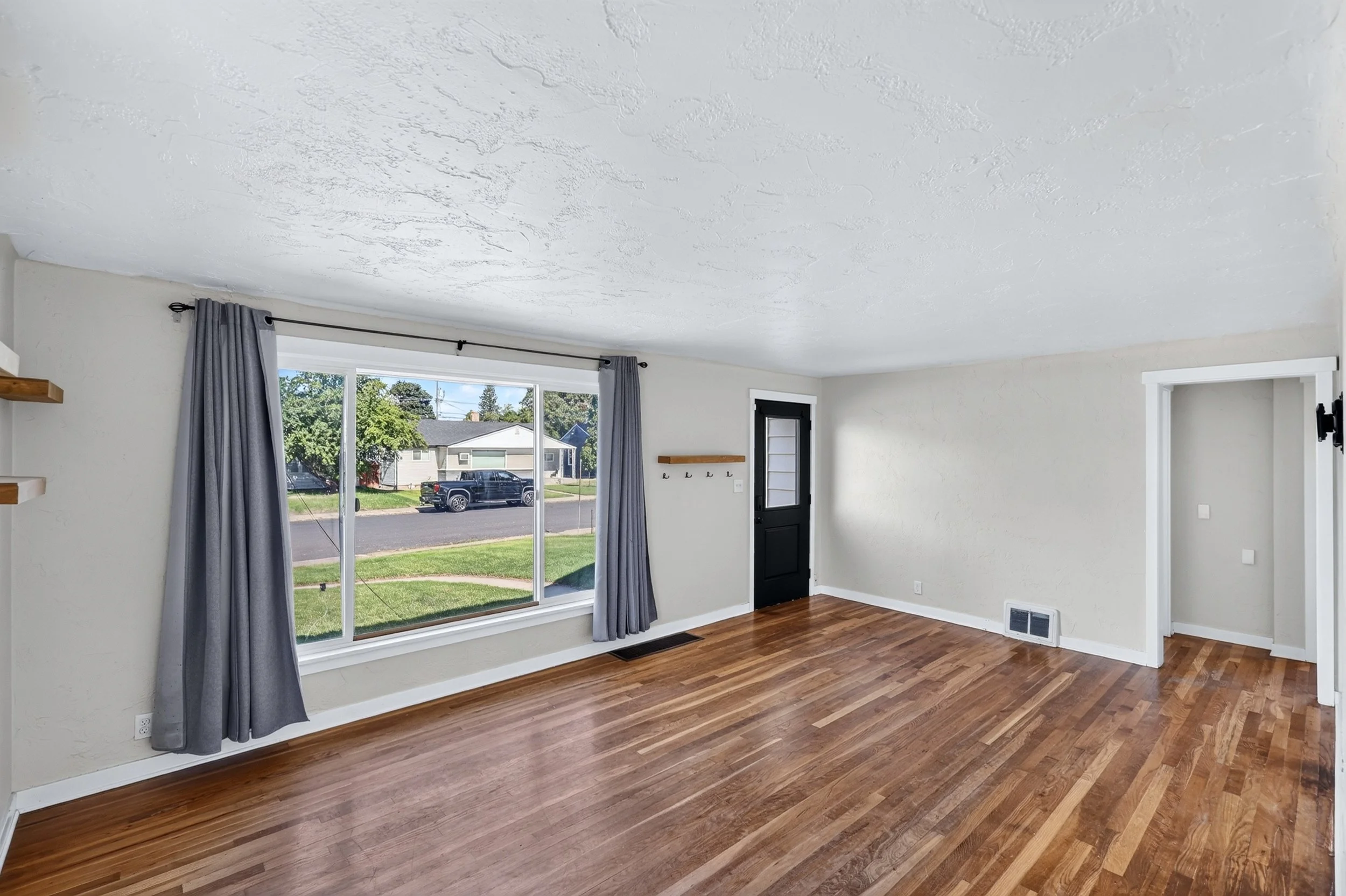 Empty living room with large window, gray curtains, hardwood floors, white walls, and a black front door.