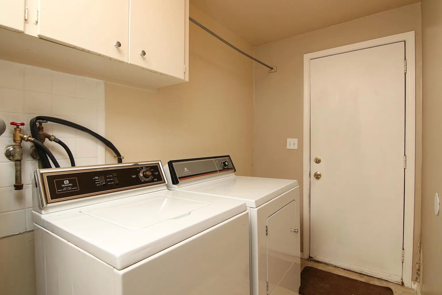 Laundry room with a white top-load washing machine and a matching dryer beside it, a white wall-mounted cabinet above, and a white door in the background.