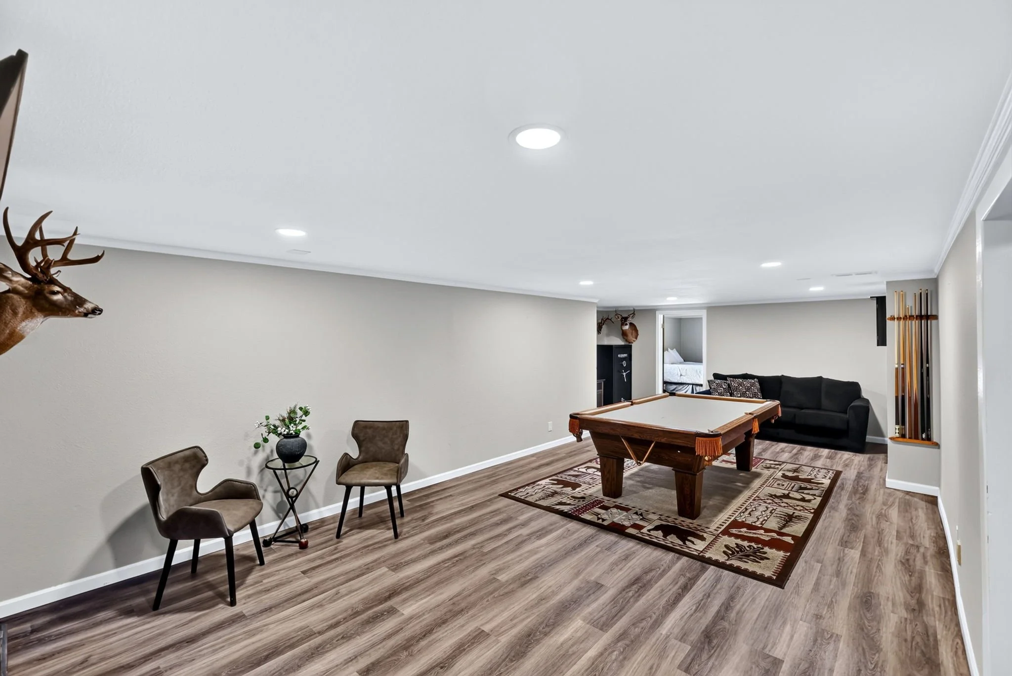 A spacious living area with beige walls and wood flooring, featuring a pool table, a black couch, mounted deer head, potted plant, and chairs, with a doorway leading to a bedroom.