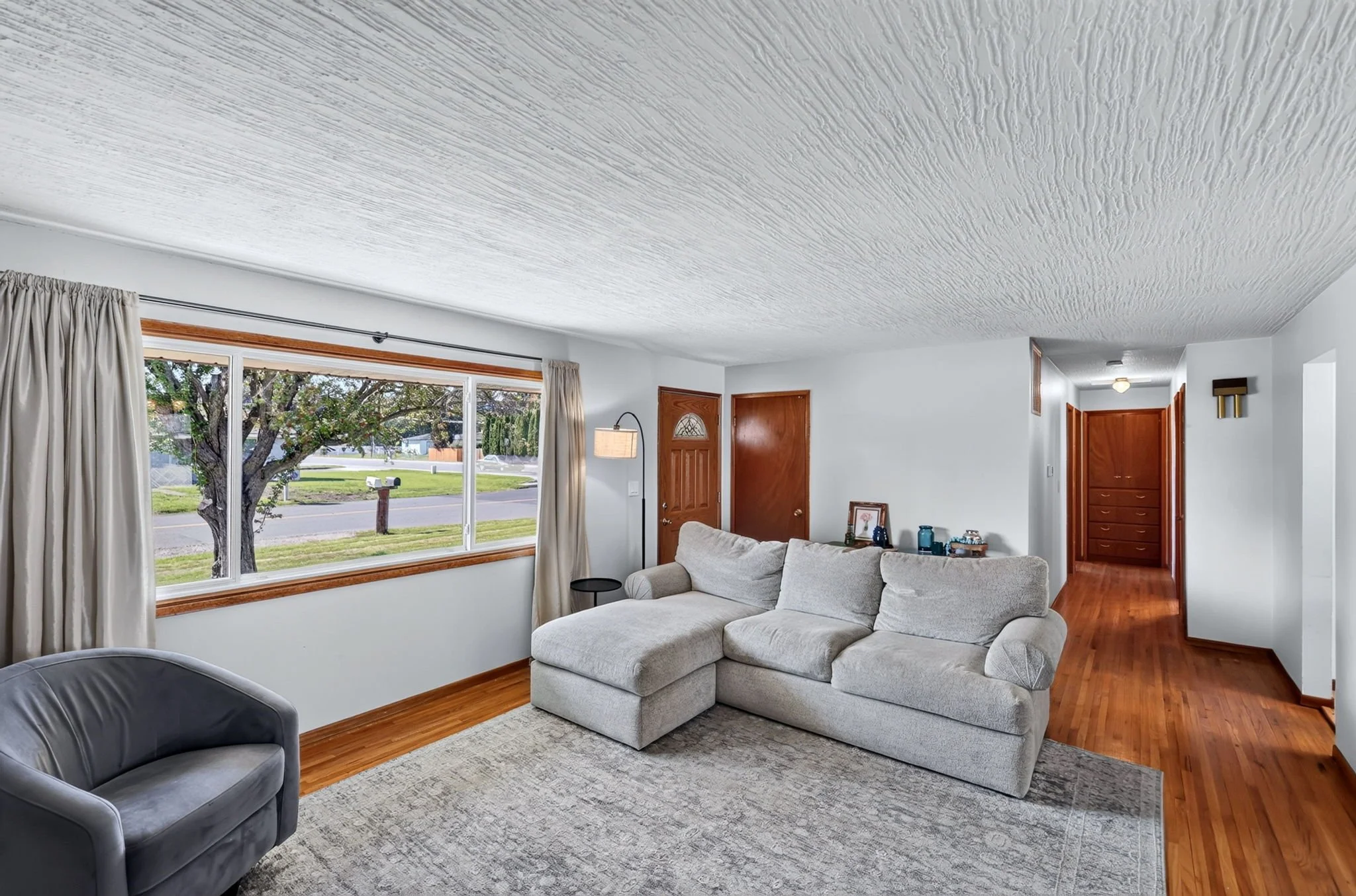 Living room with large window, white curtains, beige sofa, gray armchair, wooden flooring, and decorative items.