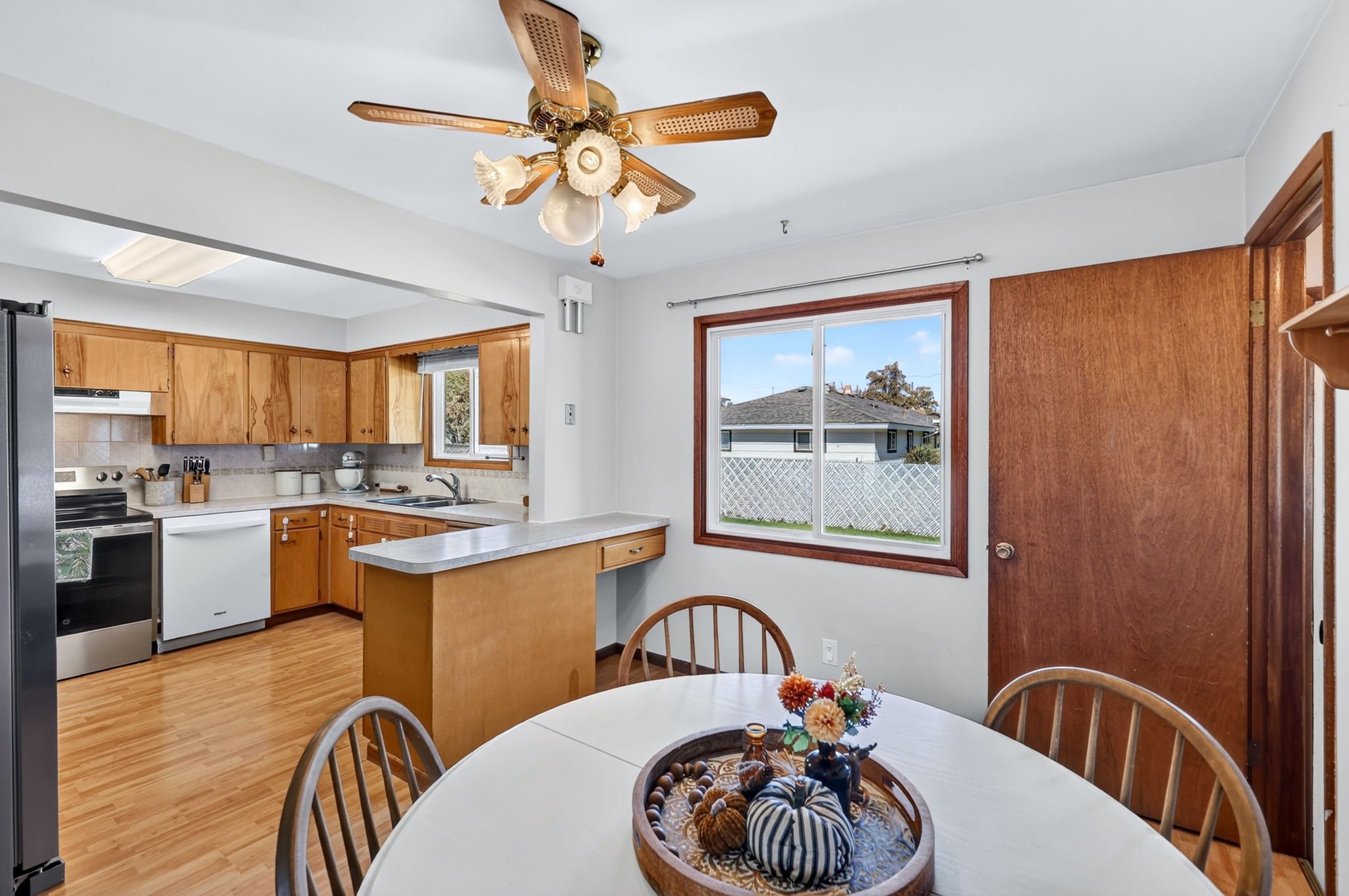 A kitchen with wooden cabinets, a white countertop, a window with a view of neighboring houses, and a ceiling fan with wooden blades and floral-style light fixtures.