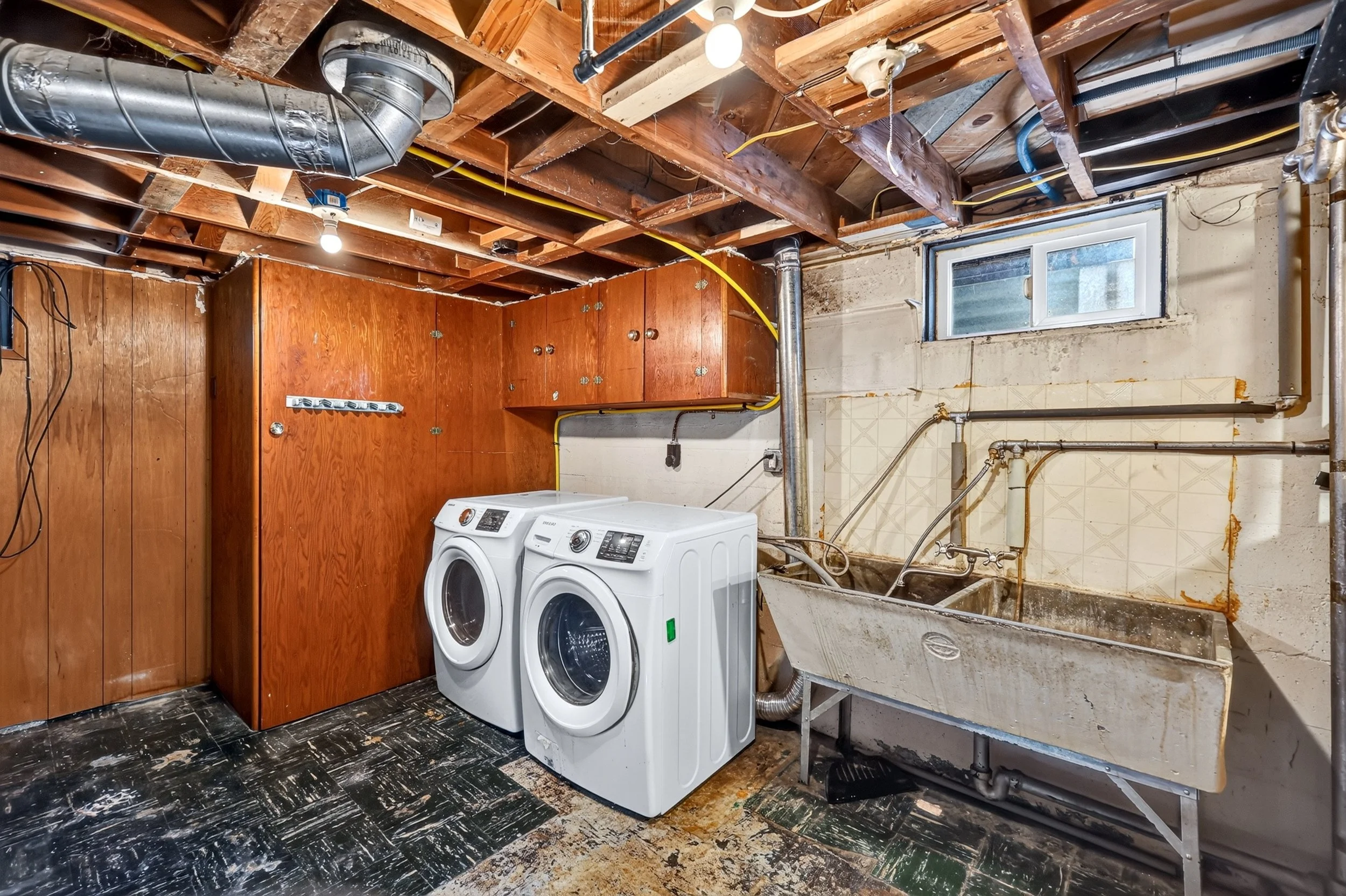 A basement laundry area with exposed ceiling beams, ductwork, and plumbing. There are two white front-loading washing machines, a wooden cabinet, a laundry sink, and a small window. The walls and floor are unfinished with visible wear and damage.